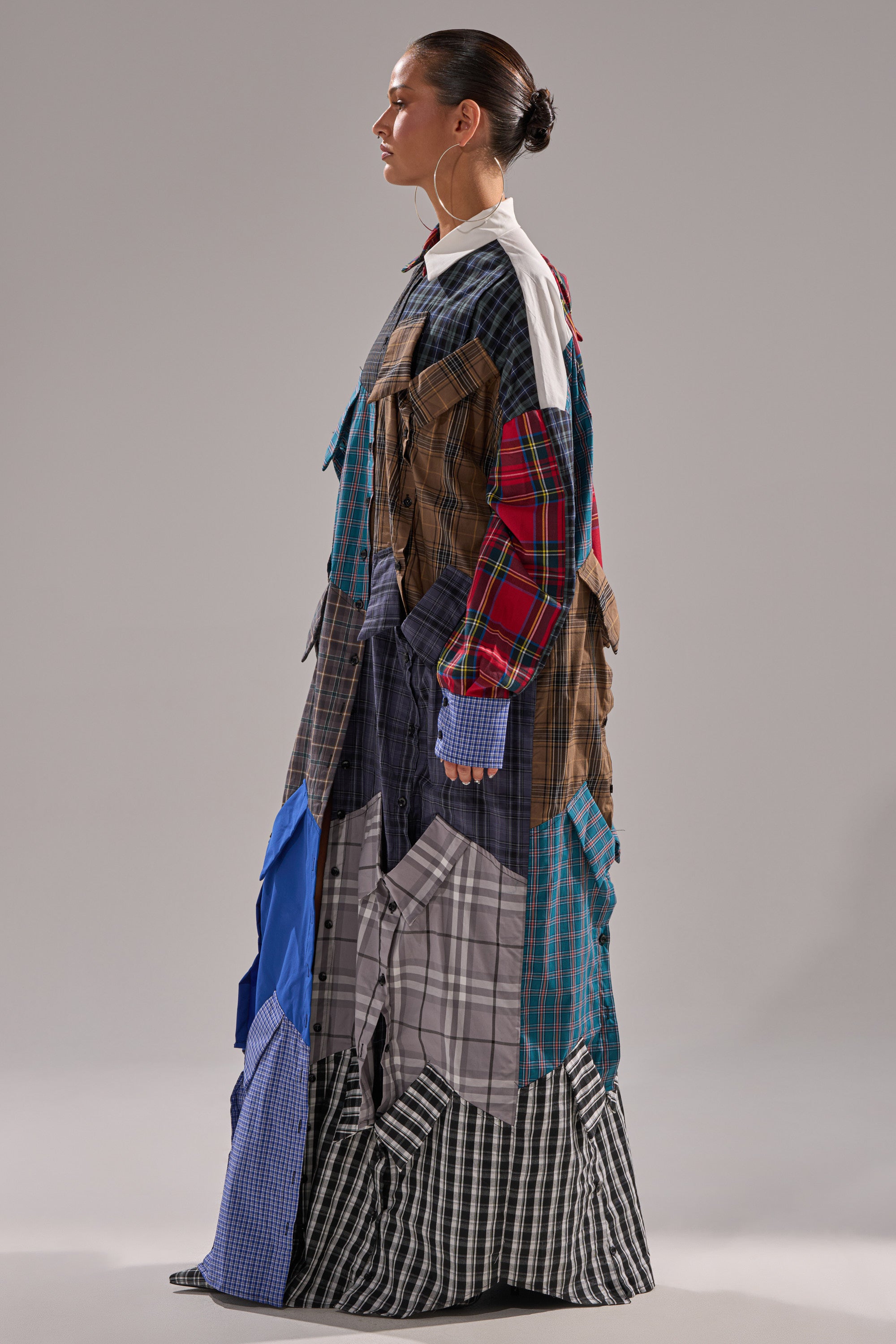 A person in profile wears the NOT YOUR BOYFRIENDS SHIRT DRESS, a long plaid shirt dress crafted from blue, red, brown, and gray checked shirts, featuring a white collar, set against a plain gray background.