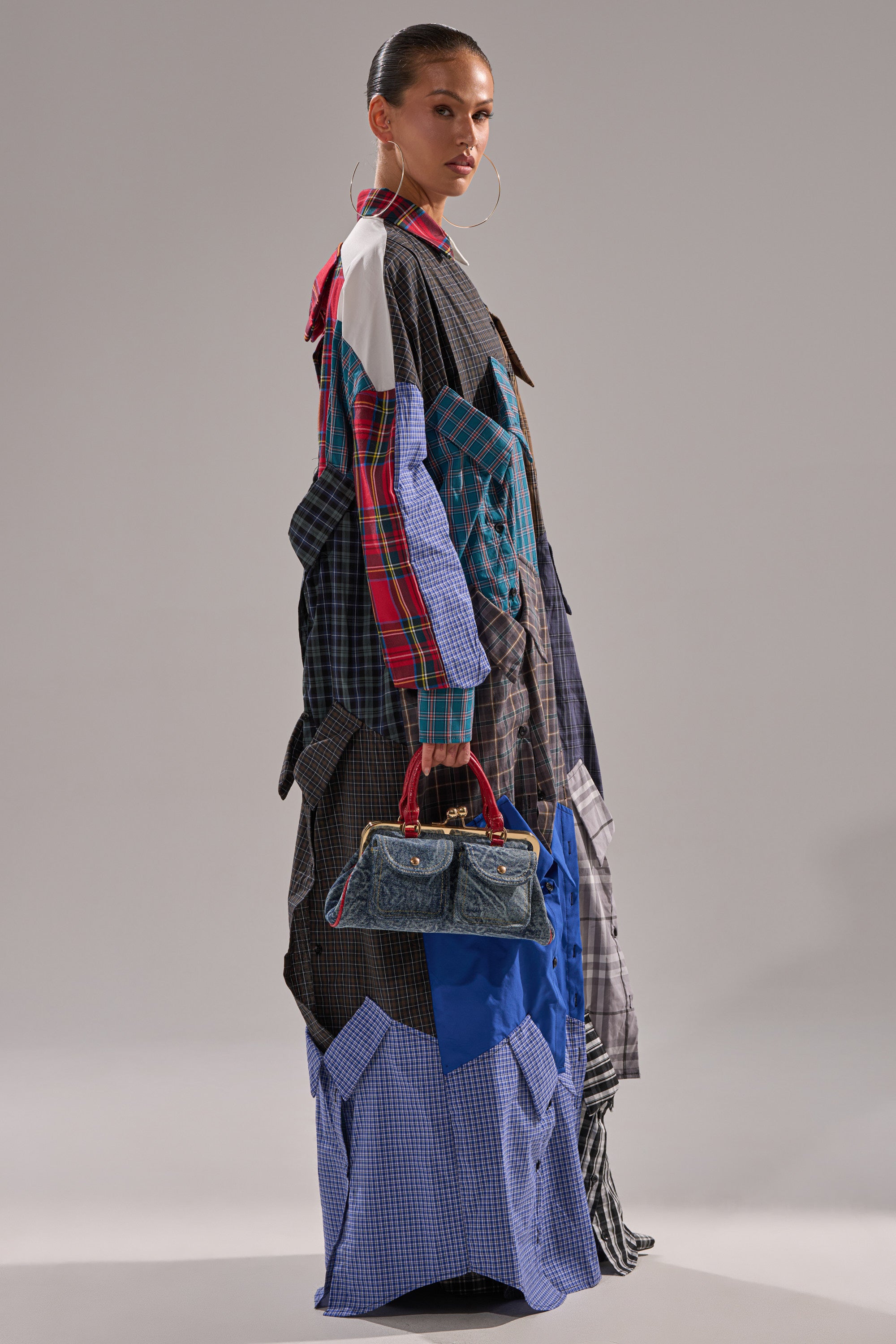 A woman stands sideways wearing the NOT YOUR BOYFRIENDS SHIRT DRESS, a vibrant plaid patchwork dress. She holds a small blue denim handbag, has slicked-back hair, and accessorizes with large hoop earrings.