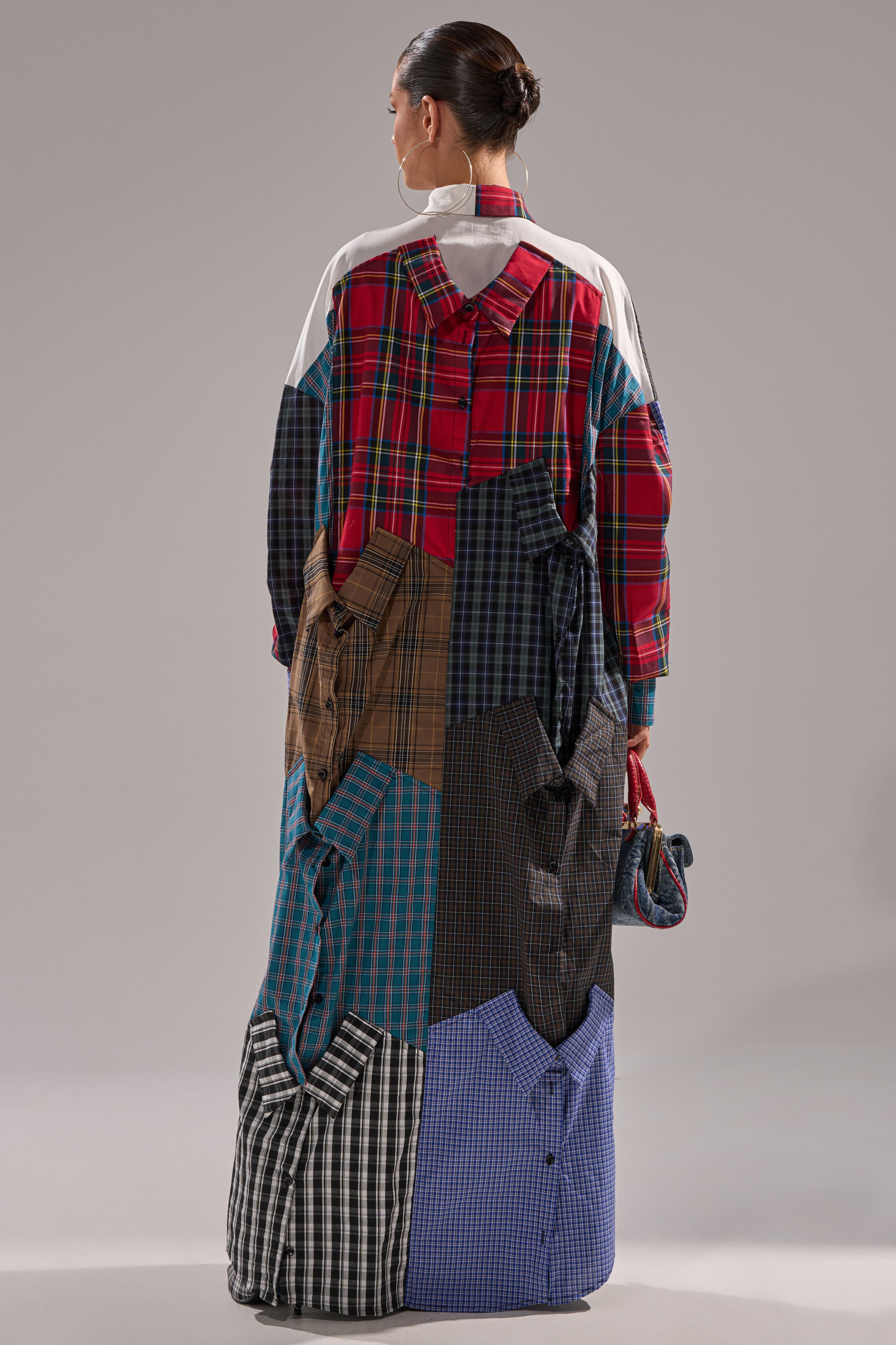 A person faces away, wearing the NOT YOUR BOYFRIENDS SHIRT DRESS—a button-down made from various colored and patterned shirts with visible collars, sleeves, and pockets. Their hair is styled in a sleek bun.