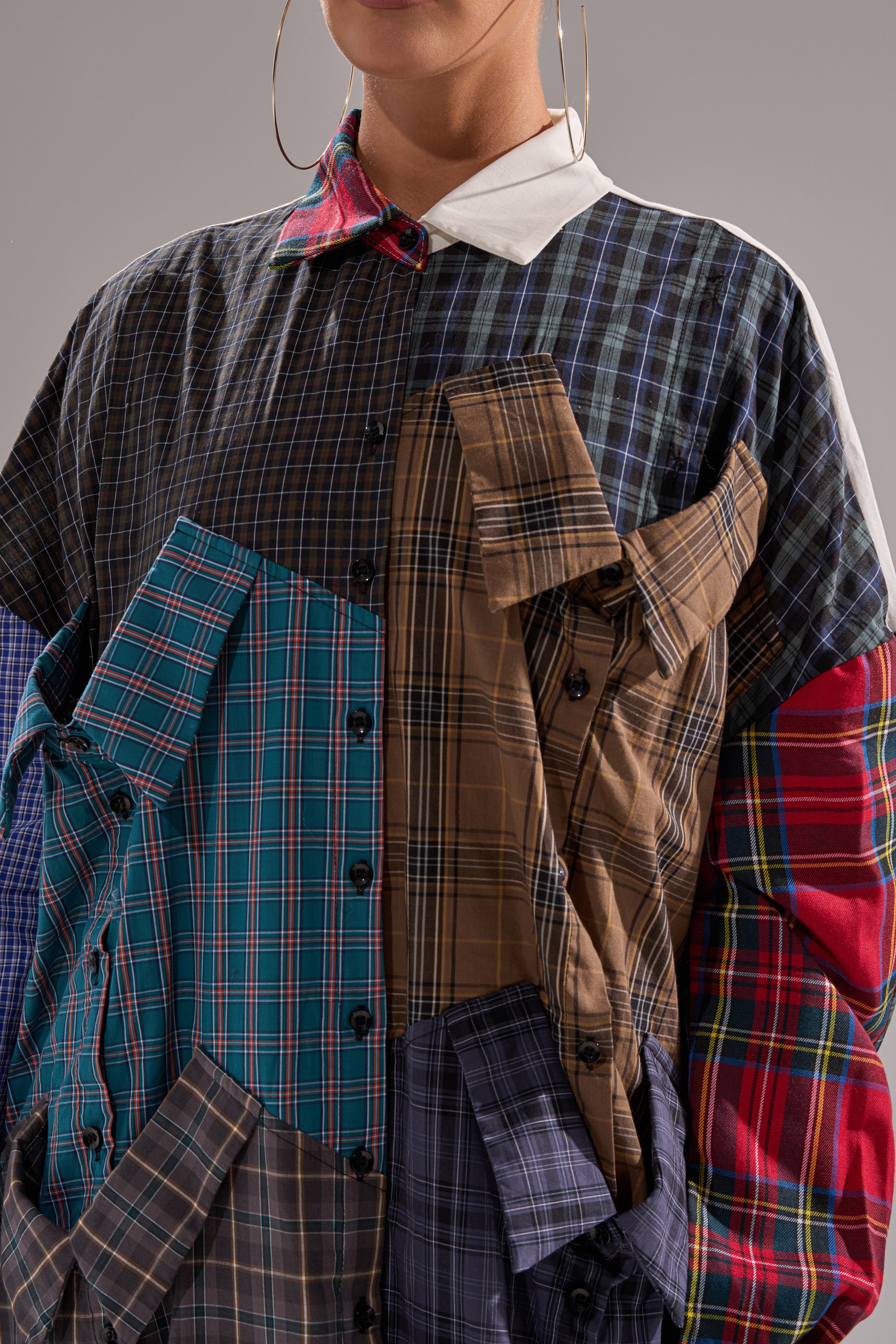 A person wearing the NOT YOUR BOYFRIENDS SHIRT DRESS, a patchwork shirt dress crafted from various colorful plaid fabrics with extra shirt fronts, pocket details, and large hoop earrings.