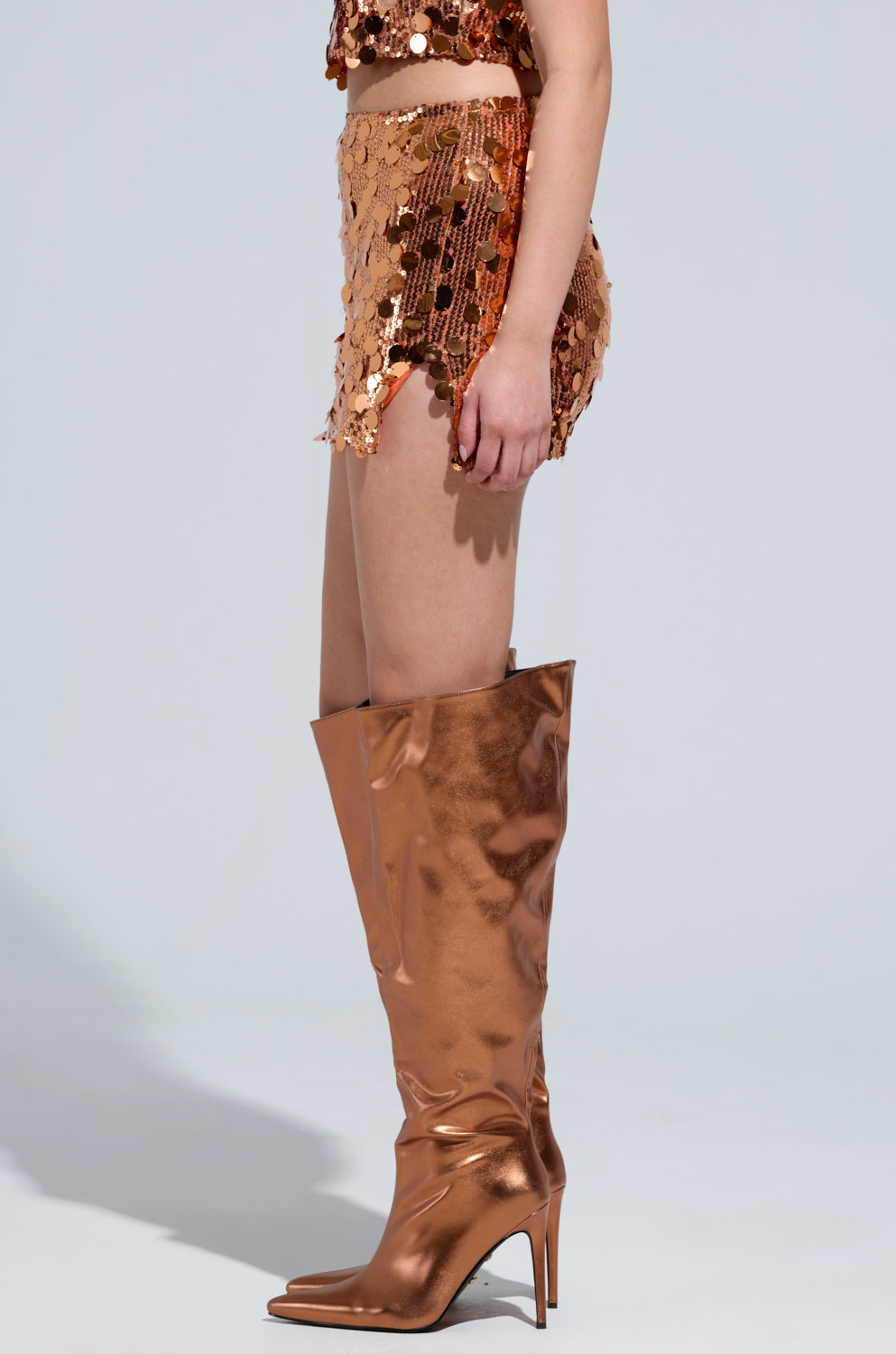 Wearing the YOURS TRULY MINI SKIRT, a person exudes Disco Diva vibes in a shimmering bronze sequin ensemble. The matching top and mini skirt are complemented by knee-high, pointed-toe bronze boots, while the plain light gray background flawlessly emphasizes the dazzling sequin discs and metallic sheen of their outfit. 2