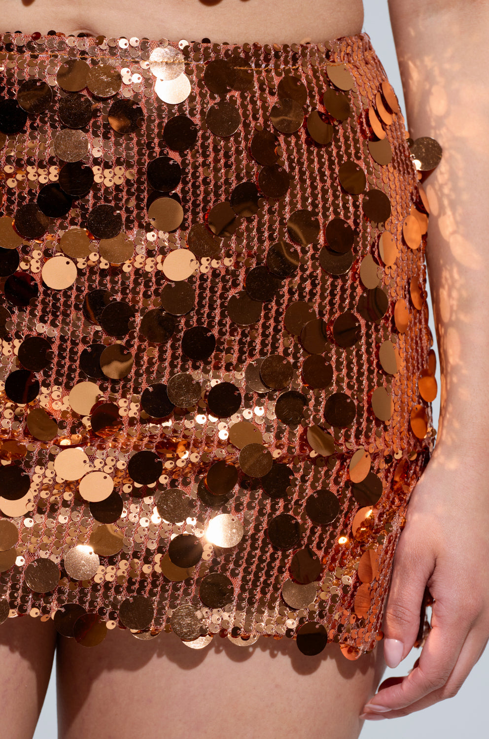 A close-up captures someone wearing the YOURS TRULY MINI SKIRT, embellished with radiant, round copper-colored sequins. Sunlight glints off the fabric, amplifying its shimmer. The person's hand is partially visible on the right side of the image, perfectly embodying a Disco Diva vibe. 6