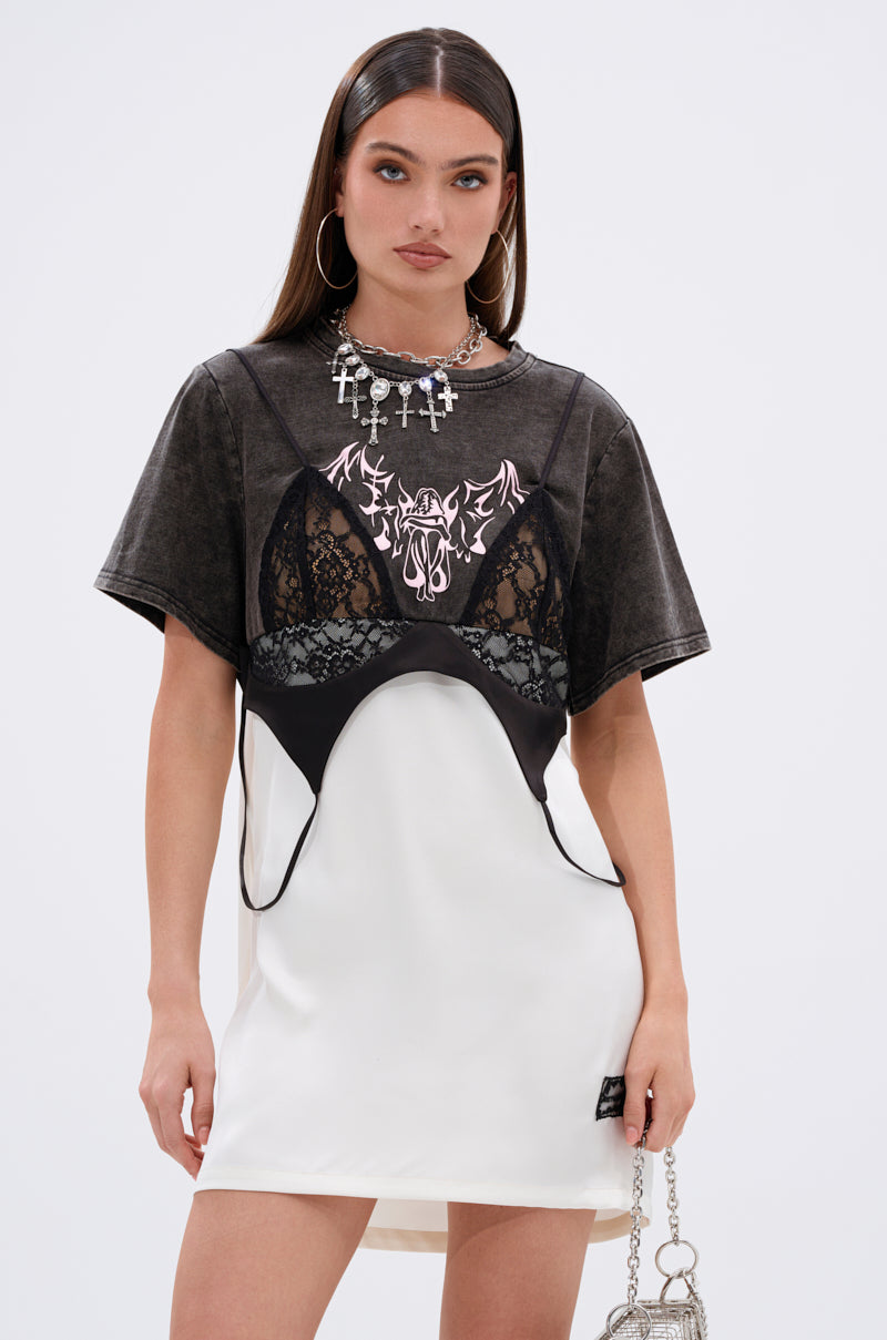 A woman with long brown hair wears the QUIT STARING GRAPHIC TSHIRT DRESS with lace paneling over a white slip dress, accessorized with hoop earrings, a silver cross necklace, and a metallic chain bag.