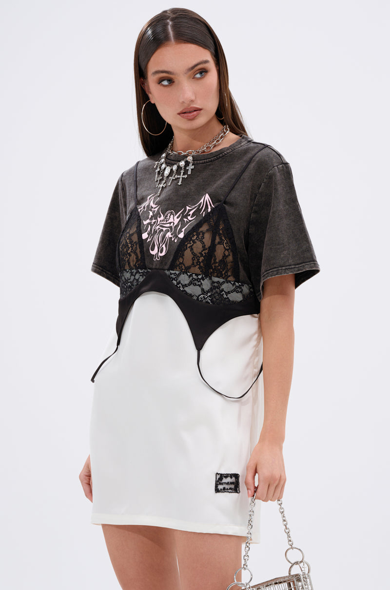 A woman styles the QUIT STARING GRAPHIC TSHIRT DRESS under a black bralette and white satin mini skirt, carrying a silver chain handbag and finishing the look with large hoop earrings, a bold necklace, and long dark hair.