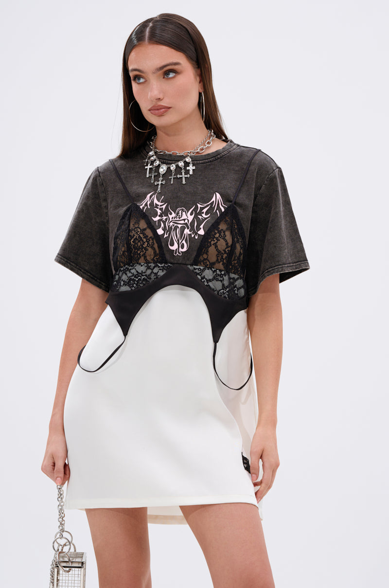 A woman wears the QUIT STARING GRAPHIC TSHIRT DRESS with lace panels over a white dress, showcasing a Gothic bat-wing illustration. She completes her look with a small silver bag and chunky cross-adorned silver jewelry.