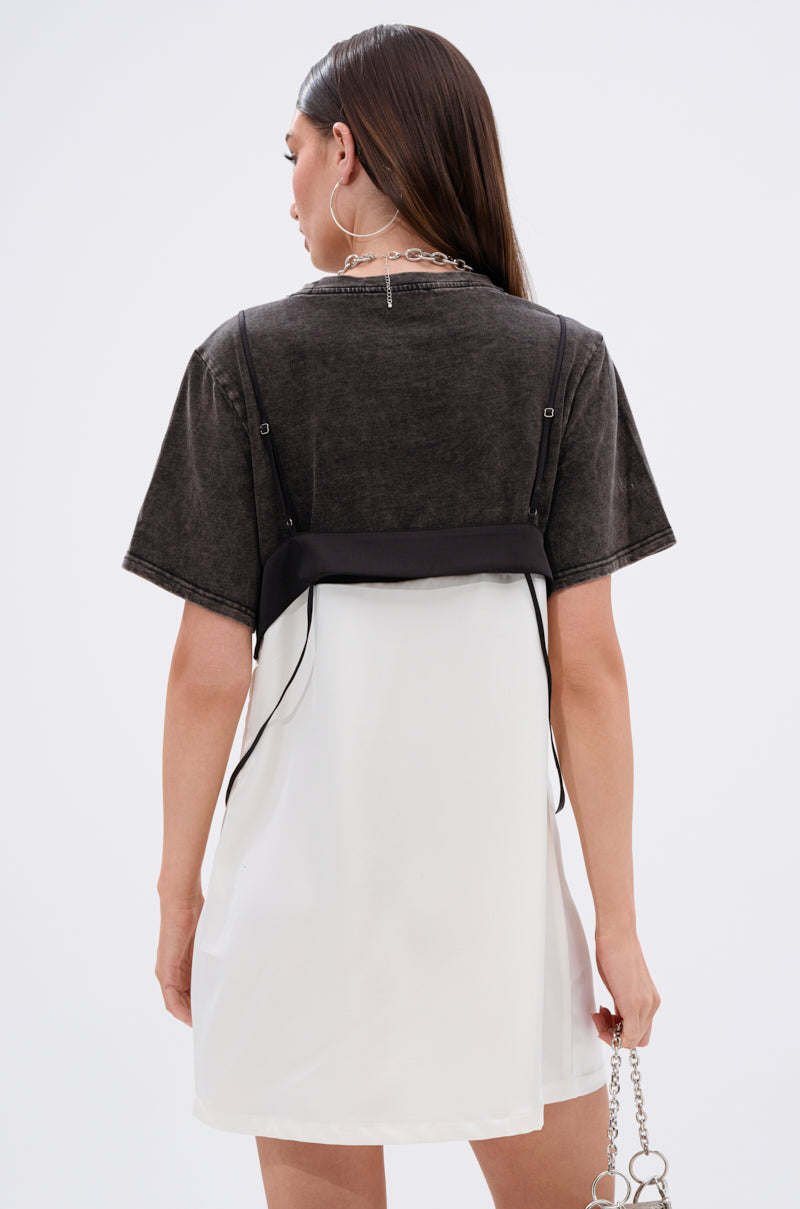 A woman with long brown hair faces away, wearing the QUIT STARING GRAPHIC TSHIRT DRESS in dark gray over a loose white dress. She holds a silver chain accessory and wears a silver necklace.