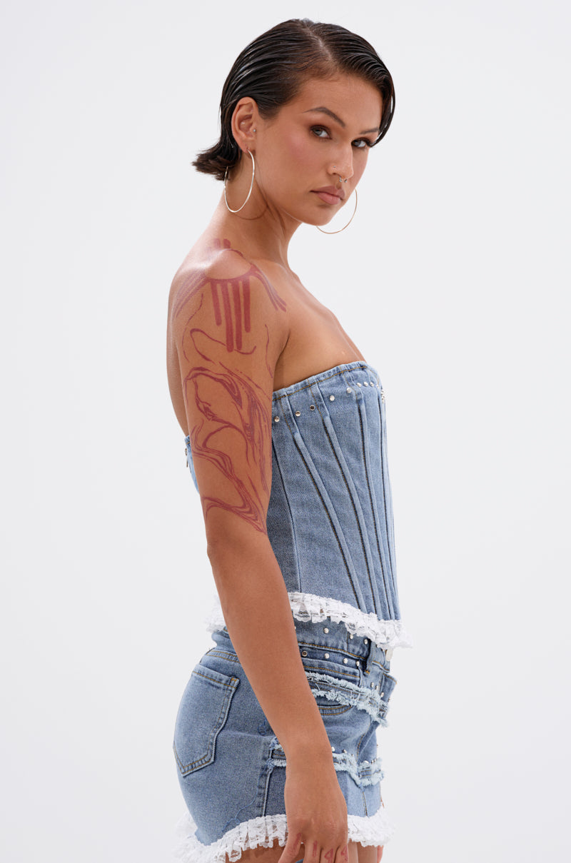 A woman with short dark hair, hoop earrings, and a red arm tattoo models the DARLING LACE TRIM DENIM CORSET—vintage blue wash with lace trim and a strapless sweetheart neckline—paired with matching denim shorts, in profile on a white background. 2