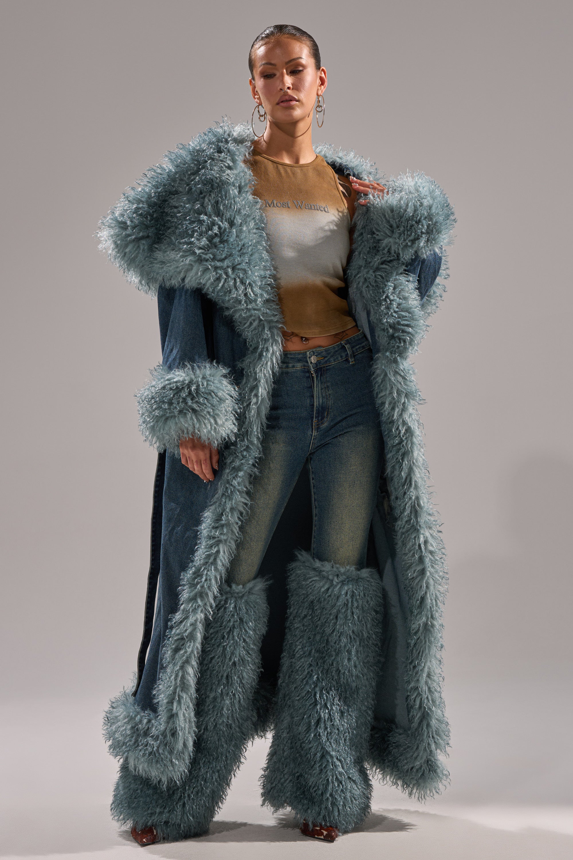 A person models the MOXIE FUR TRIM DENIM TRENCH with faux fur leg warmers, jeans, a brown T-shirt, and large hoop earrings, standing against a plain background.