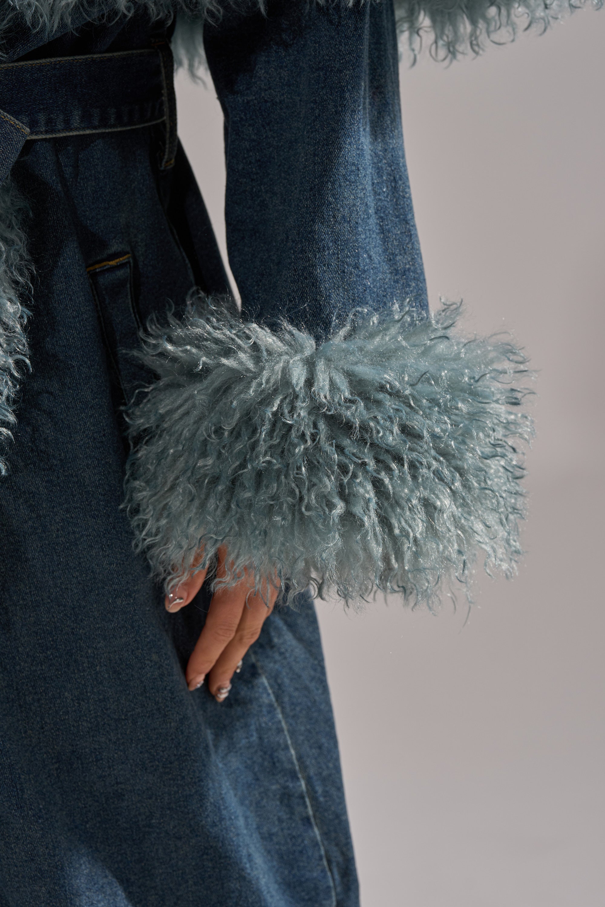 A close-up shows a person wearing the MOXIE FUR TRIM DENIM TRENCH, featuring fluffy light blue fur-trim cuffs. Their hand rests beside the coat against a plain light background.