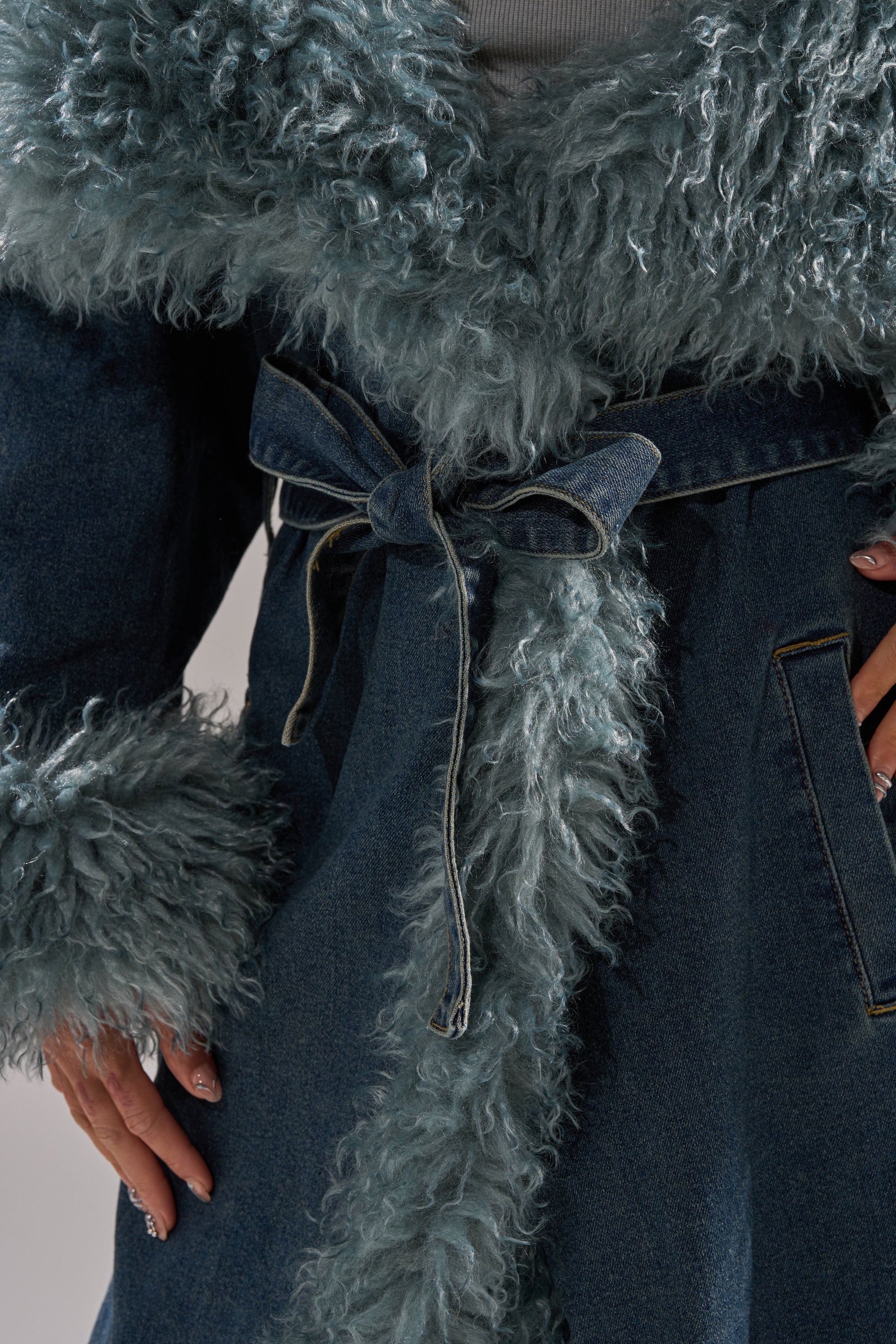 A close-up of a person wearing the MOXIE FUR TRIM DENIM TRENCH with fluffy fur trim and a matching denim belt tied at the waist, hands resting on the sides of the longline jacket.