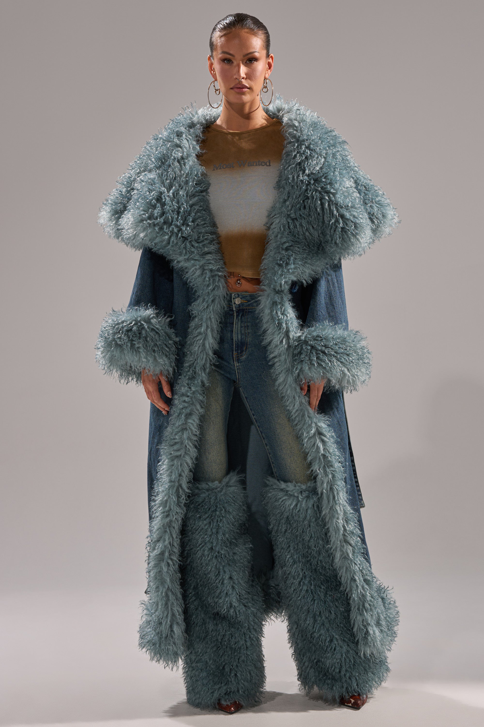 A woman faces forward, wearing the MOXIE FUR TRIM DENIM TRENCH with oversized pale blue fur on the collar, cuffs, and hem, paired with matching denim jeans and large pale blue fur leg warmers.