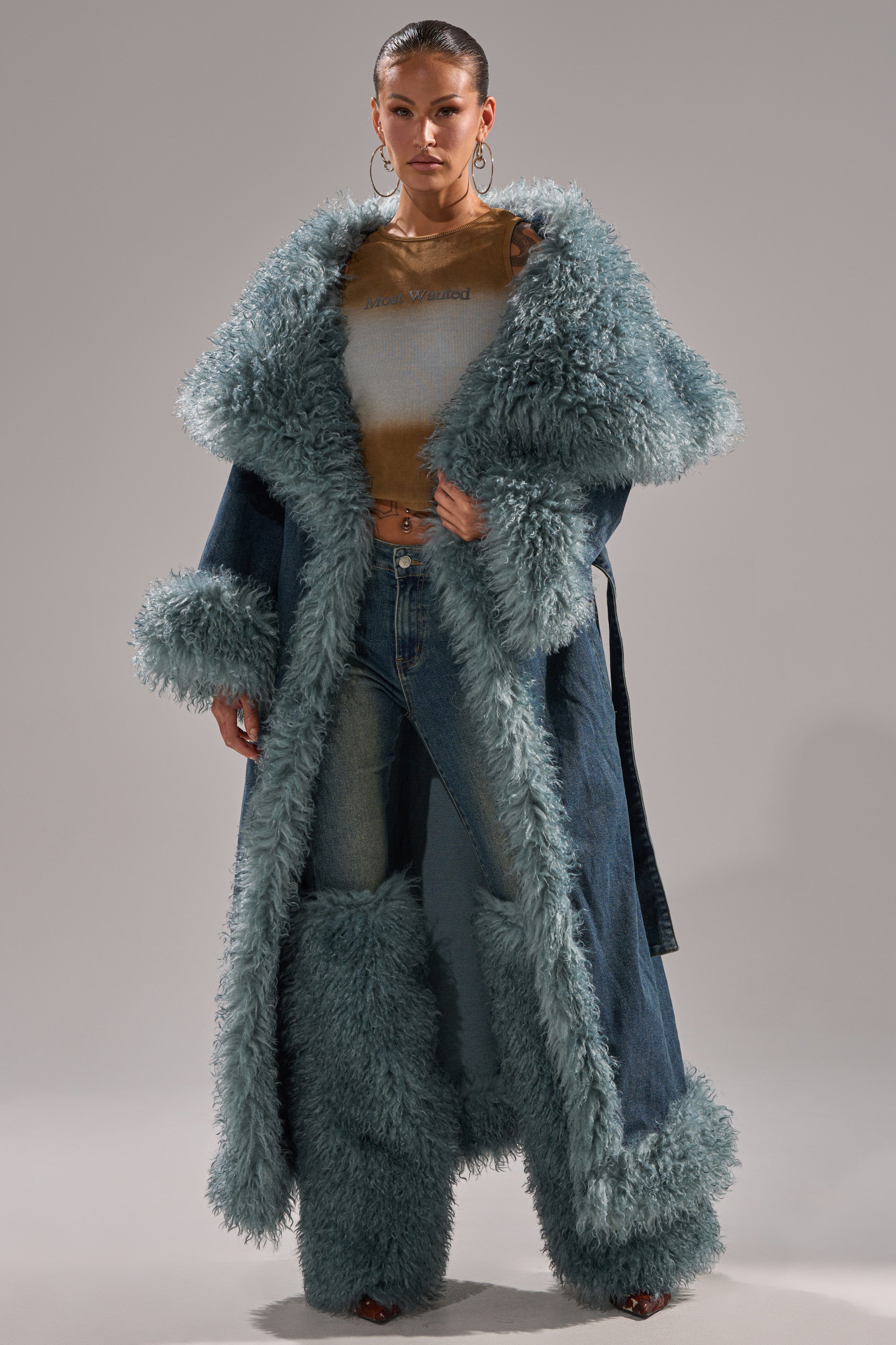 A woman poses against a gray background wearing the MOXIE FUR TRIM DENIM TRENCH with thick pale blue fur trim, matching fur leg warmers, light blue jeans, a brown sheer top, and large hoop earrings.