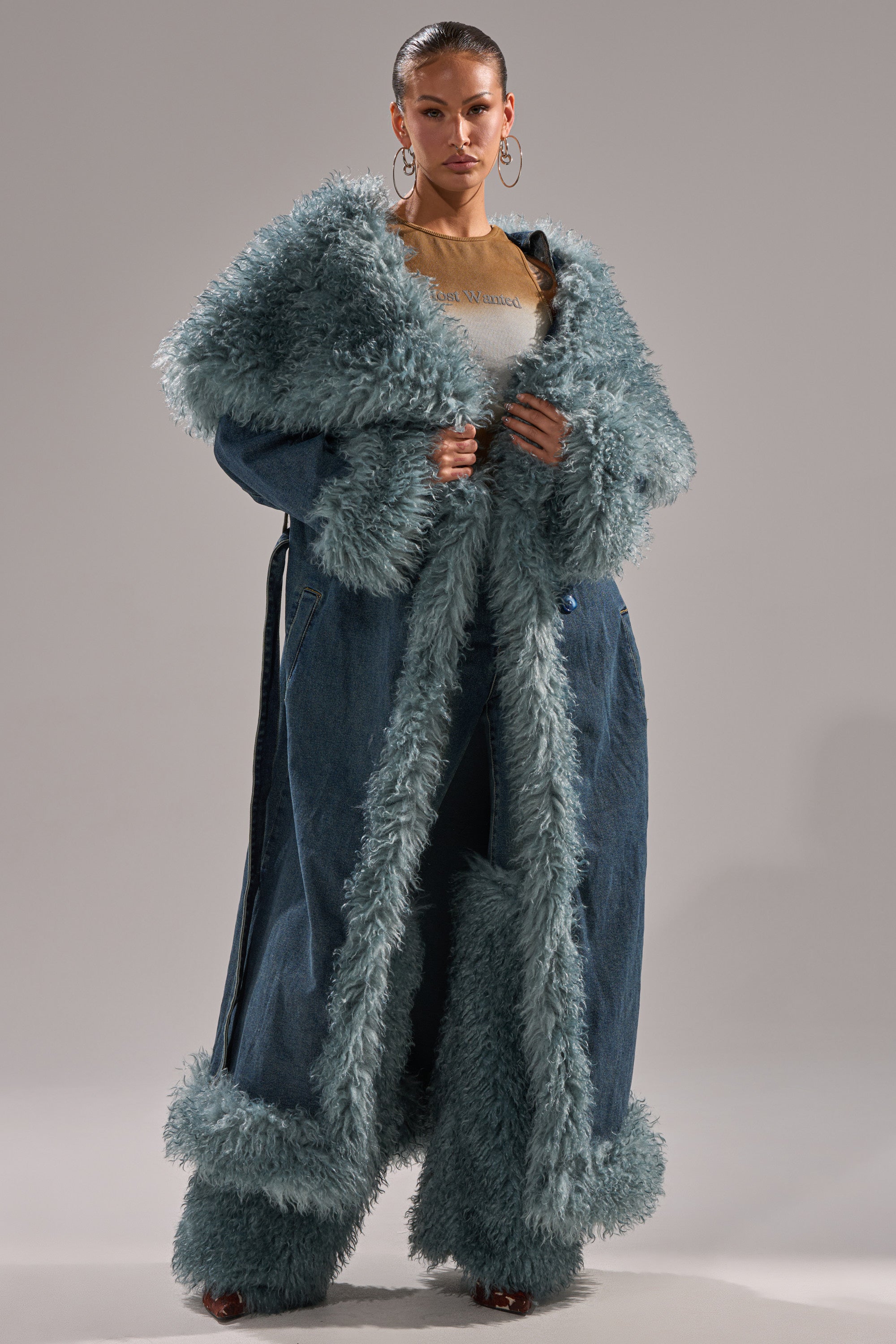 A woman wears the MOXIE FUR TRIM DENIM TRENCH, featuring longline denim with fluffy light blue fur on the collar, cuffs, hem, and front edge. She accessorizes with hoop earrings and poses confidently against a plain background.