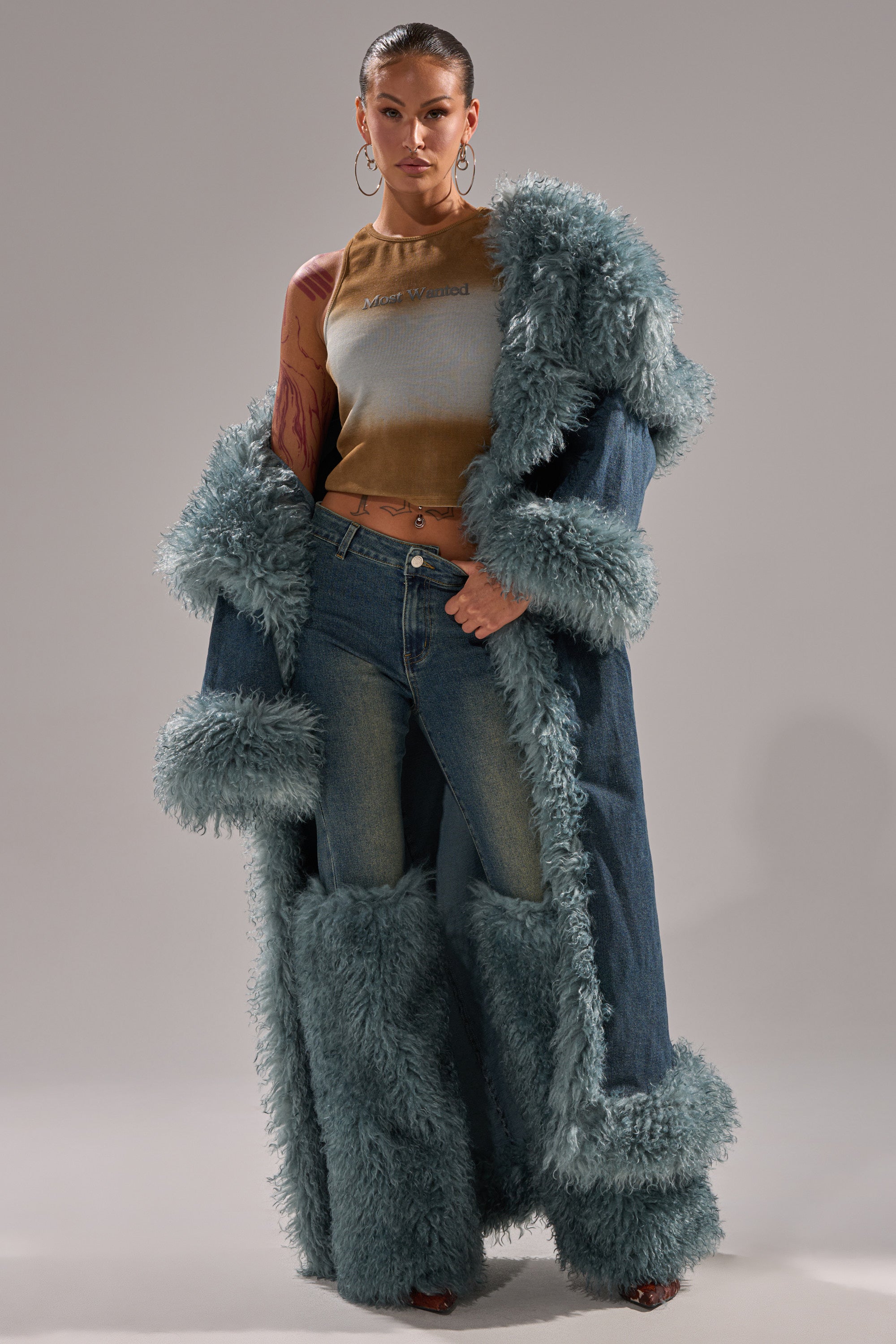 A woman poses confidently in the MOXIE FUR TRIM DENIM TRENCH with fluffy fur trim and matching jeans, styled with a tan sleeveless top and hoop earrings, against a plain gray background.