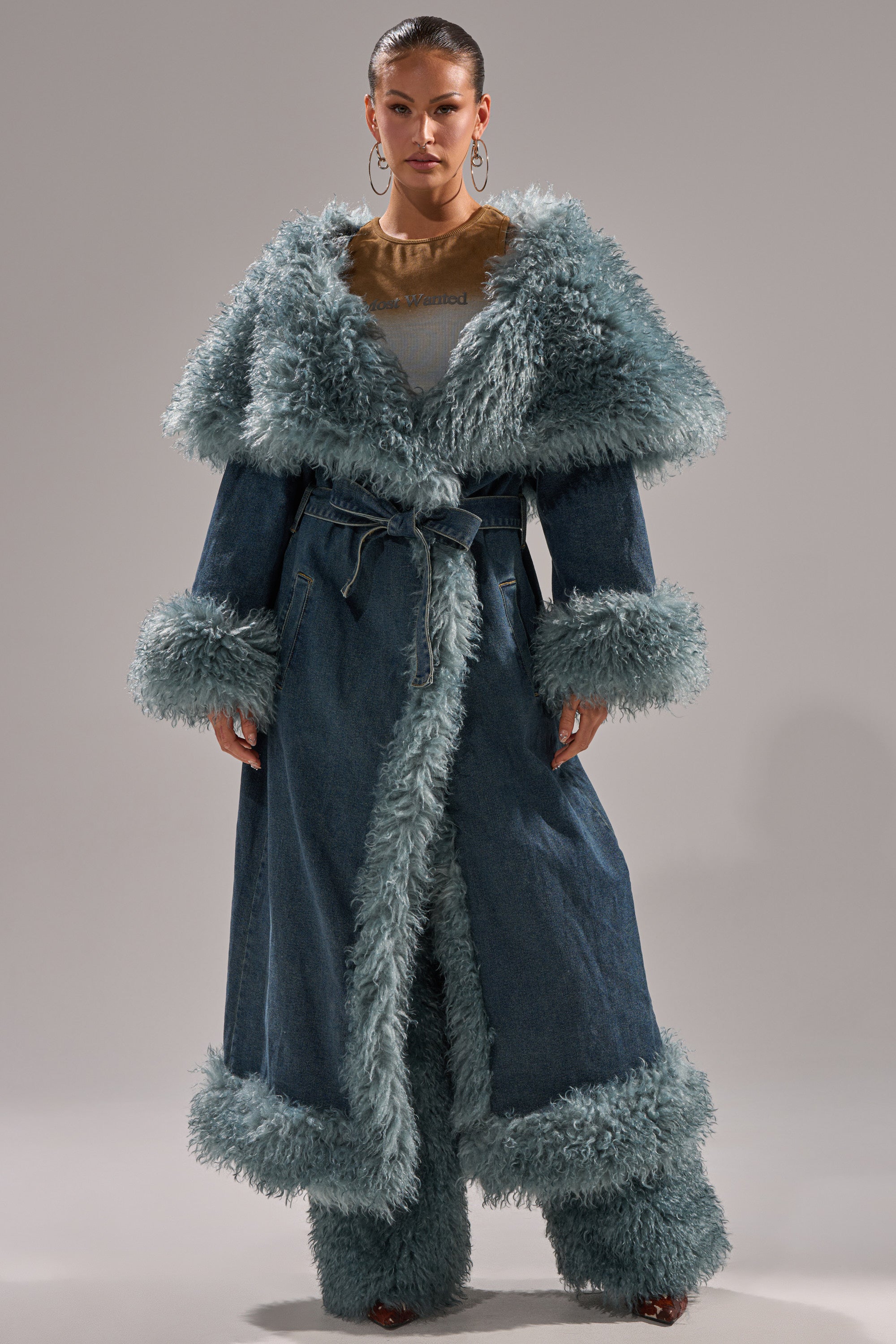 A woman faces forward in the MOXIE FUR TRIM DENIM TRENCH, featuring large light blue fur trim on the collar, cuffs, and hem. She pairs it with matching fur-trimmed denim pants and hoop earrings against a plain gray background.