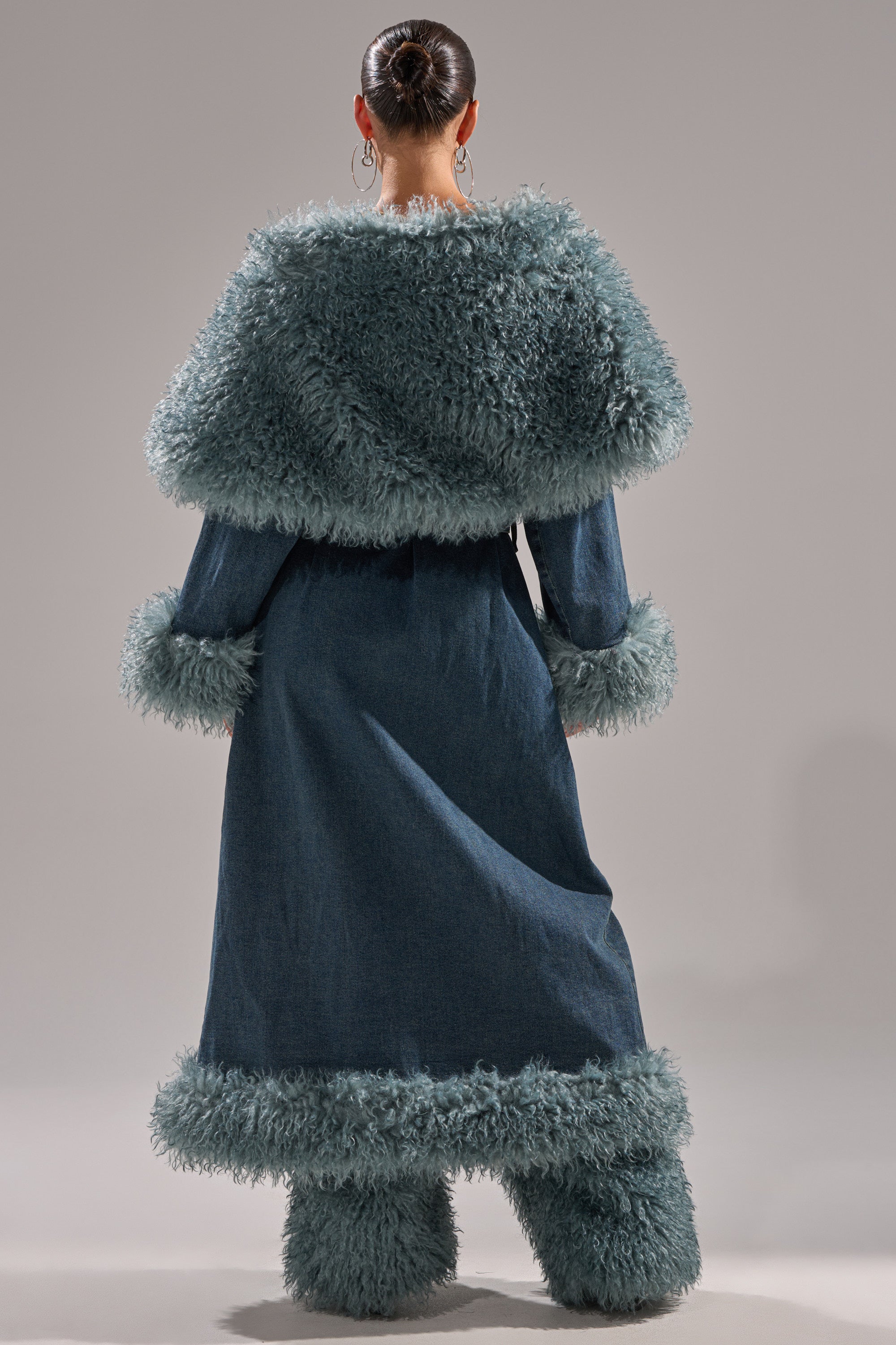 A person with hair in a neat bun wears the MOXIE FUR TRIM DENIM TRENCH, featuring blue denim with fur trim on the collar, sleeves, and hem, paired with matching boots. They stand against a plain light gray background.