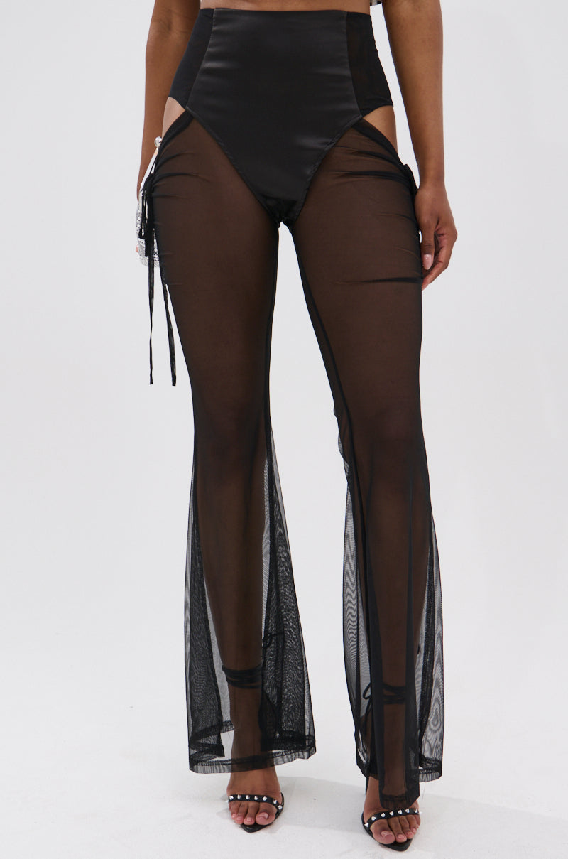 A person wears DERRIA MESH FLARE LEGGING, high-waisted and sheer with ruched drawstring sides, high-cut hips, and open-toe heels. The mesh pants reveal the legs and tie at the hip, set against a plain white background. 1