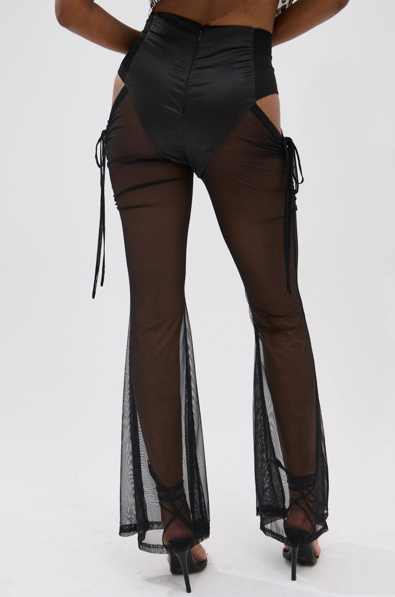 Model wearing DERRIA MESH FLARE LEGGING—high-waisted black mesh flares with ruched drawstrings, cut-out sides, and high-cut hips—paired with black heels, photographed from the back against a plain light background. 4