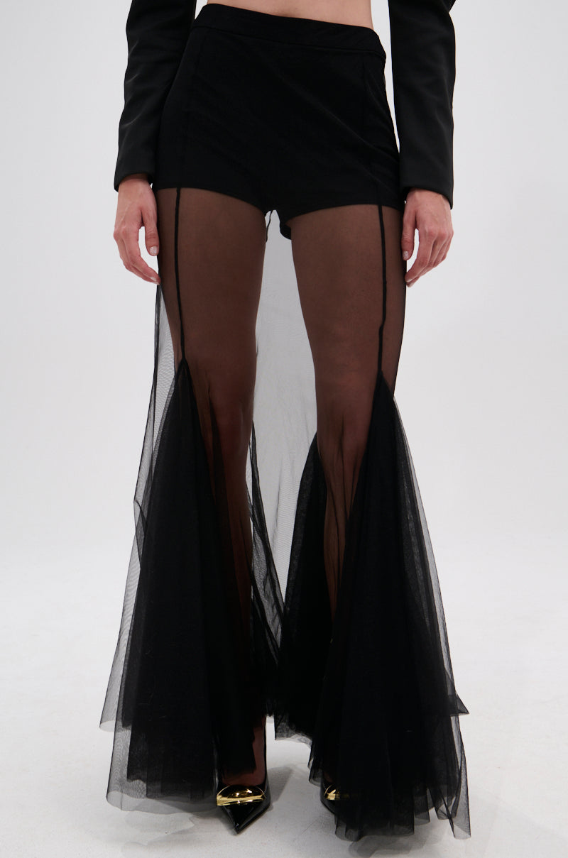 Wearing the CHLOE MESH FLARE MAXI SKIRT—high-waisted black shorts with sheer, pleated mesh maxi skirt overlays flaring at the ankles—paired with a black crop top and pointed black heels.