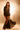 A woman stands confidently in dramatic lighting, wearing the CHLOE MESH FLARE MAXI SKIRT with a black blazer, lace-up boots, and a small round purse. Her wavy brown hair and bold look exude high-fashion style.