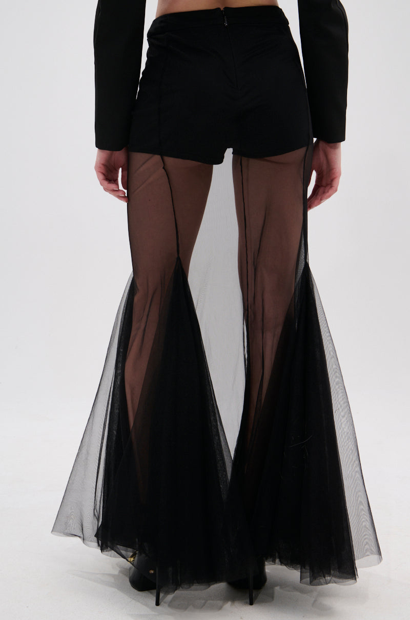 A person wears black shorts layered with the CHLOE MESH FLARE MAXI SKIRT, which features a high-rise waist and pleated mesh flare, photographed from the back against a plain light background.