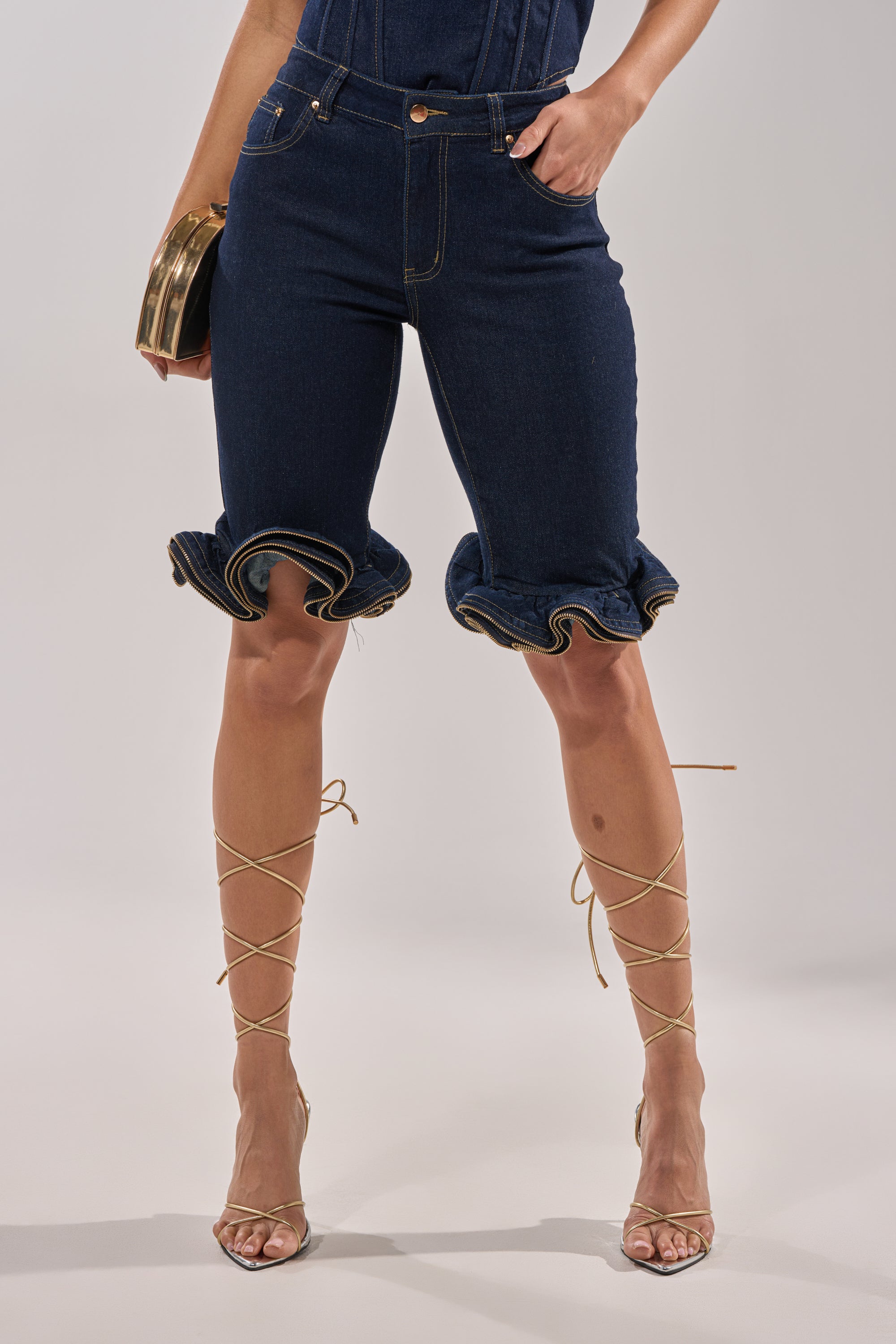 A person wears the SOMETHING DIFFERENT DENIM BERMUDA SHORT, featuring a high-rise fit, dark wash, and ruffle hems. Only the lower body is visible as they hold a clutch and stand in strappy lace-up heeled sandals against a plain background.