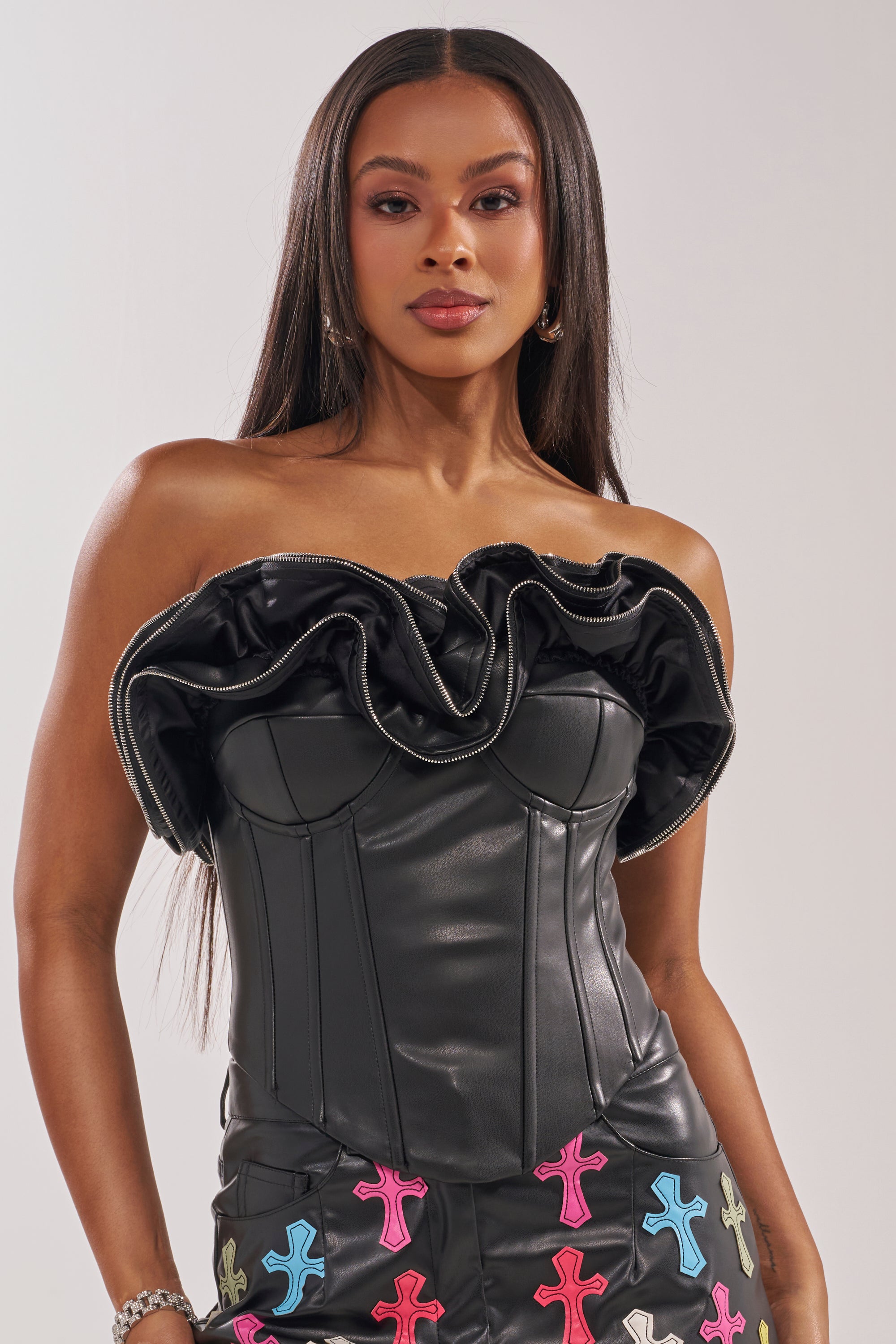 A woman with long straight hair wears the DON'T MESS RUFFLE CORSET, a strapless top with zippered ruffle detailing, paired with a matching skirt adorned with colorful cross designs, standing confidently against a plain light background. 1
