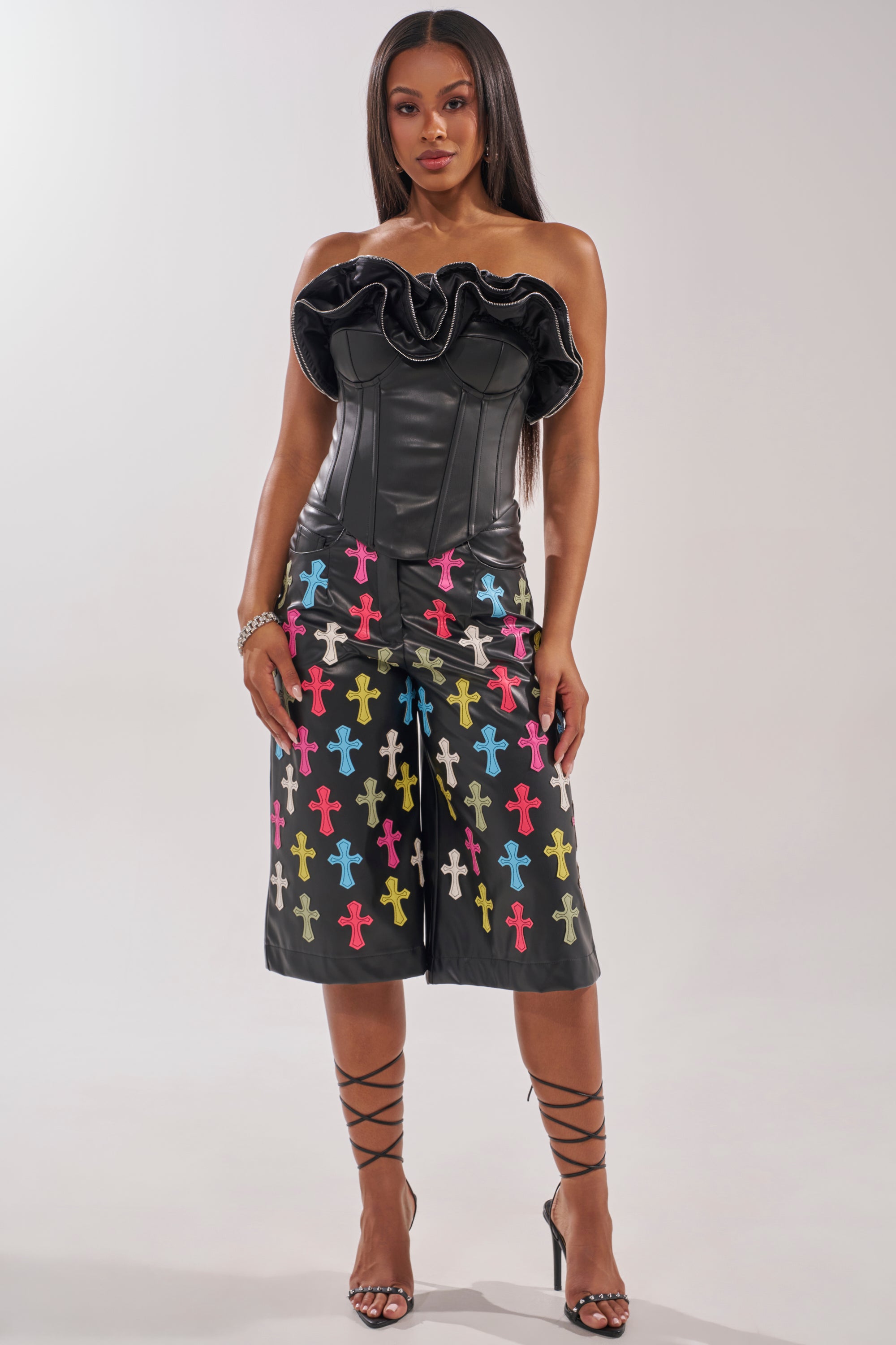 A woman wears the DON'T MESS RUFFLE CORSET, a black strapless top with ruffled zippered details, paired with matching knee-length shorts featuring colorful cross patterns. She has long straight hair, strappy heels, and a neutral expression. 4