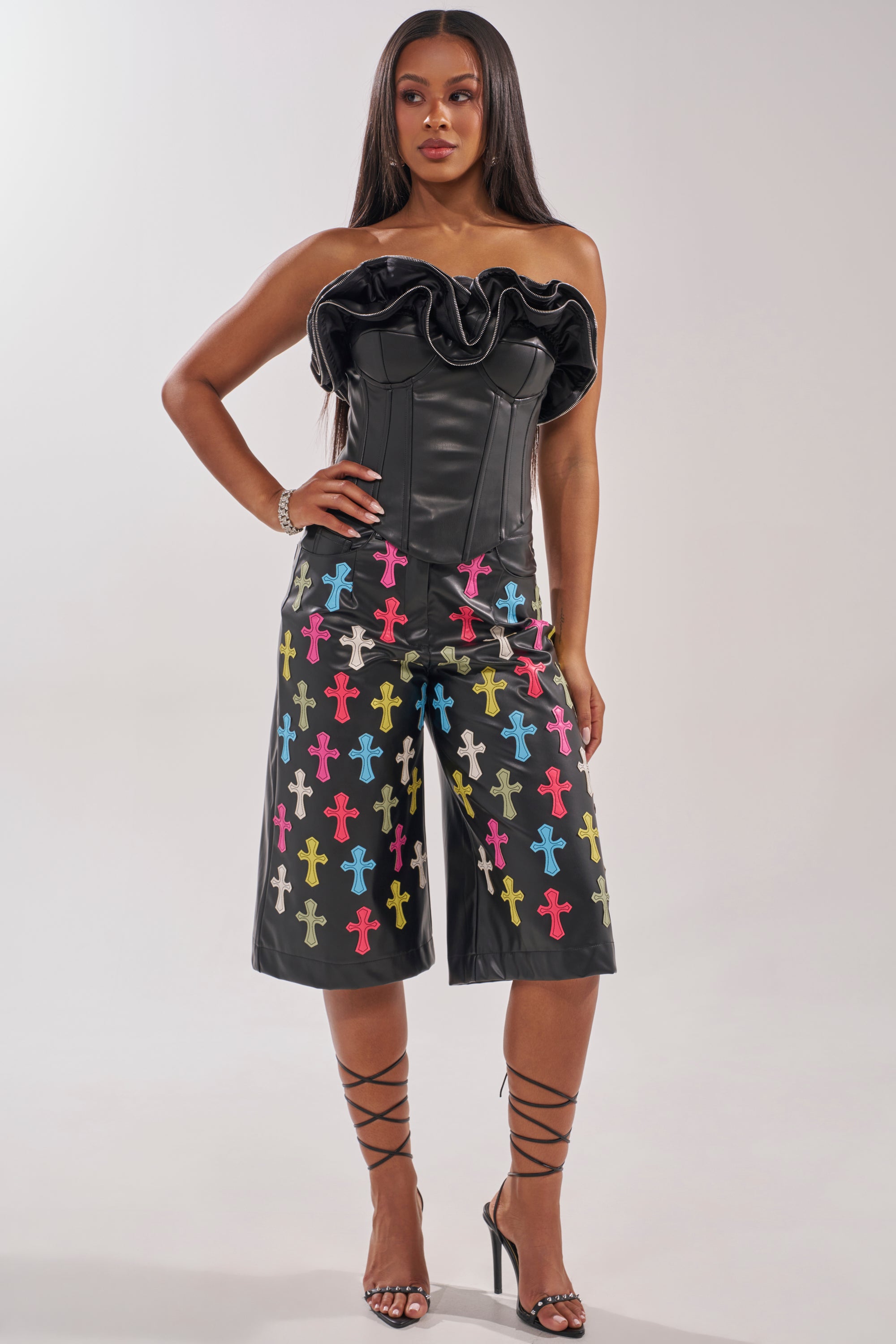A woman stands confidently in the DON'T MESS RUFFLE CORSET, a strapless leather top with zippered ruffle details, paired with black knee-length shorts featuring colorful cross patterns. She has straight hair and wears strappy black heels. 5