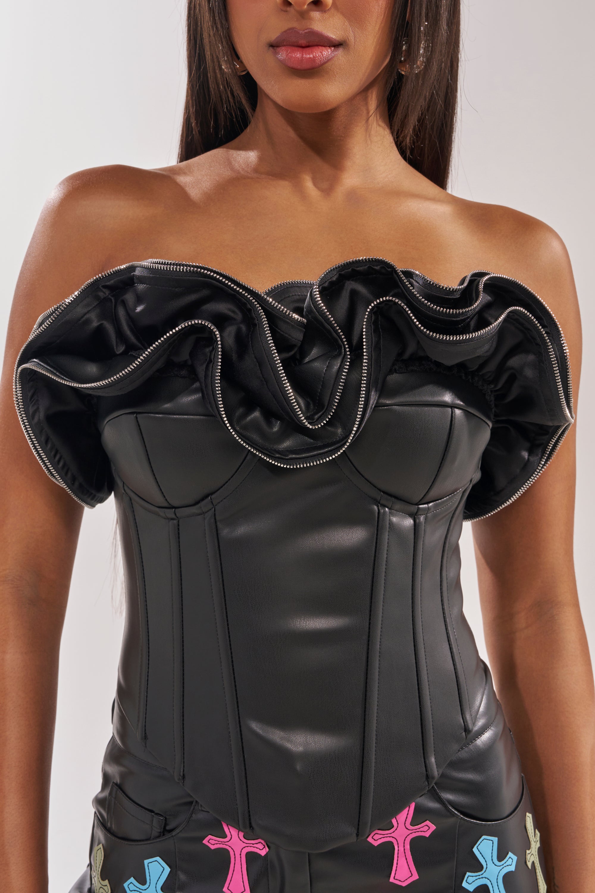 A woman models the DON'T MESS RUFFLE CORSET, a black strapless leather top with zippered ruffle trim on the neckline, paired with black pants decorated with colorful cross patches. Only her torso and part of her face are visible. 6