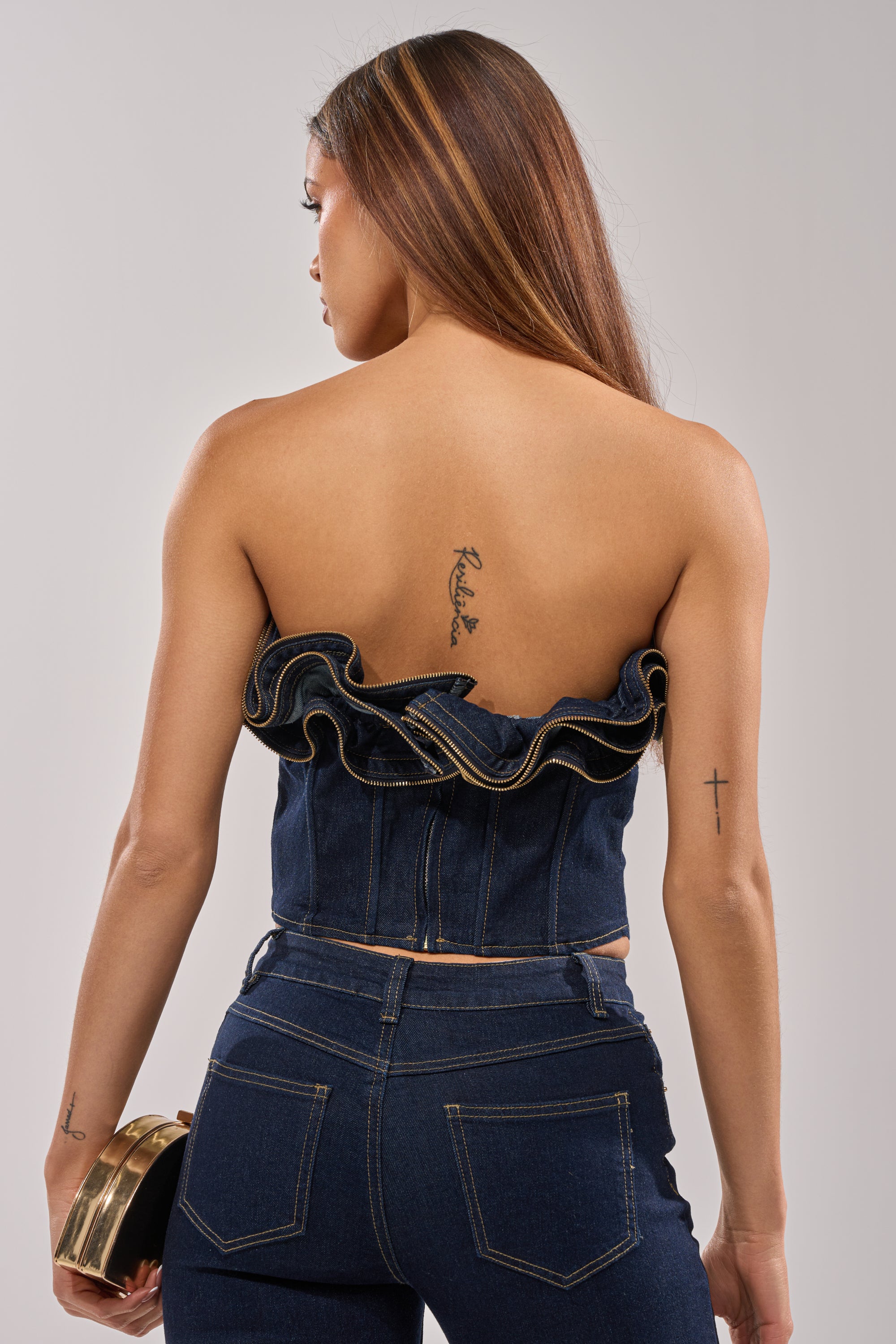 A woman with long brown hair, seen from behind, wears the SOMETHING DIFFERENT DENIM CORSET WITH ZIPPER DETAIL paired with matching jeans. She holds a small clutch and has tattoos on her back and right arm.