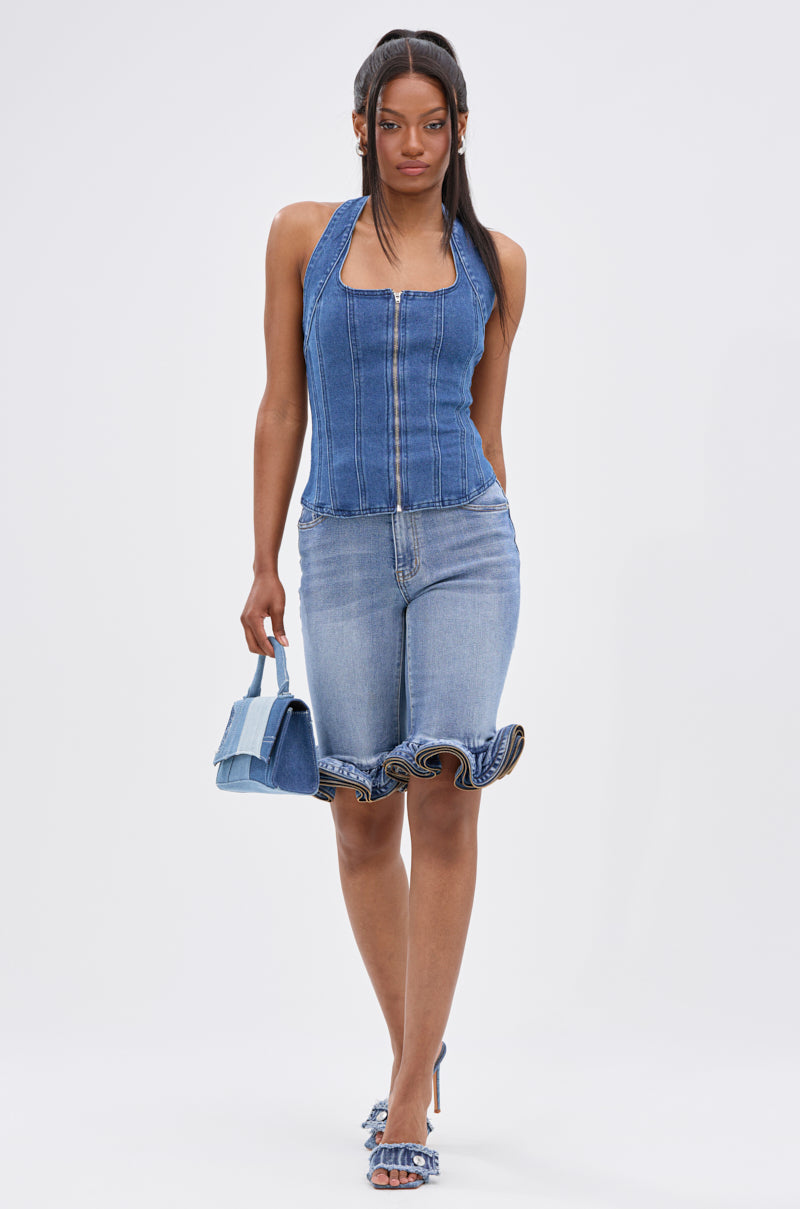 A woman wearing the LONDON BERMUDA SHORT denim bottoms walks forward, carrying a small denim handbag, styled with blue heels and long, straight hair in a half-up, half-down look.