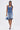 A woman wearing the LONDON BERMUDA SHORT denim bottoms walks forward, carrying a small denim handbag, styled with blue heels and long, straight hair in a half-up, half-down look.