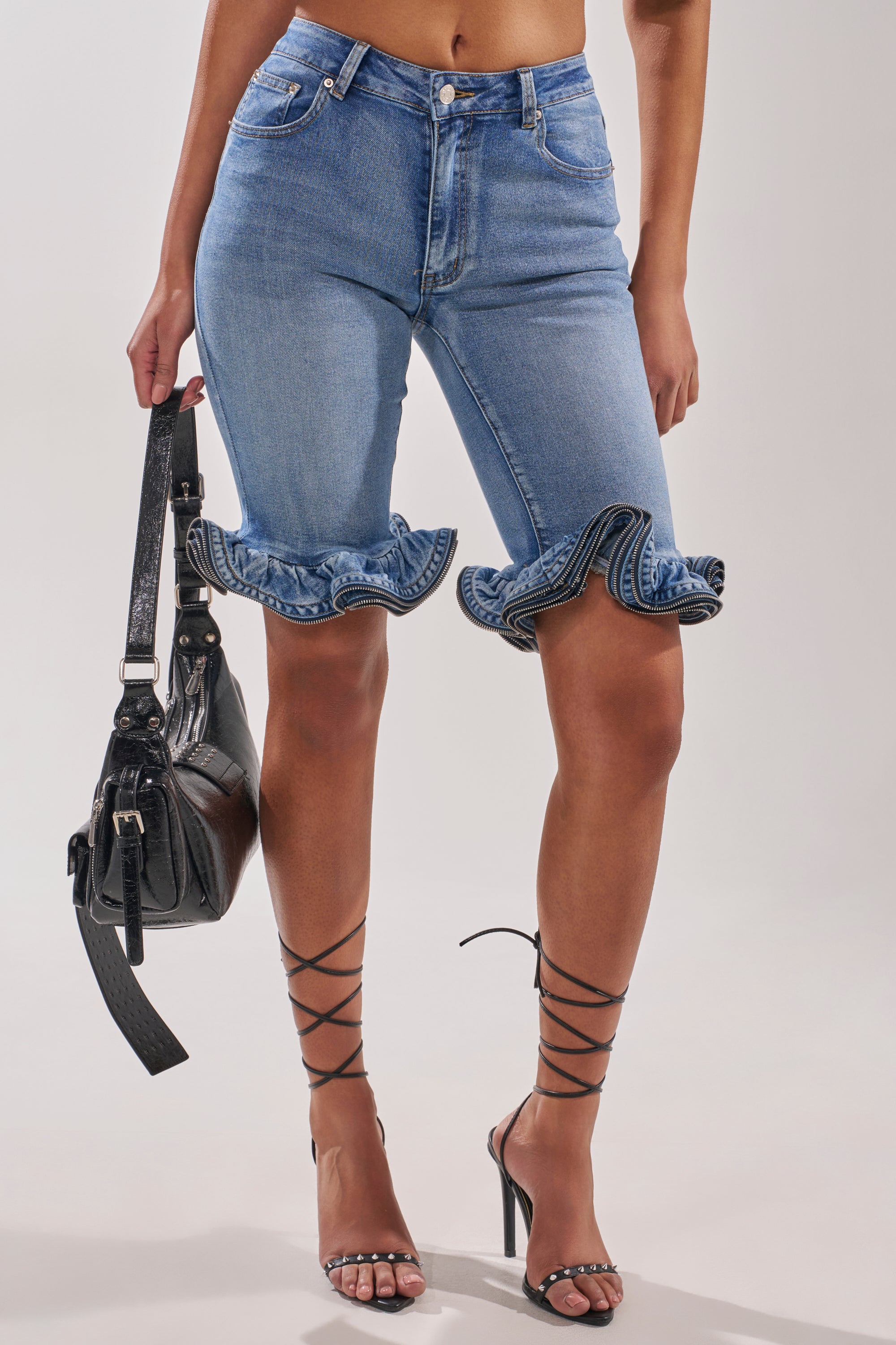 A woman wears the LONDON BERMUDA SHORT, high-waisted denim shorts with a ruffled hem above the knees, styled with strappy black heels and a matching black handbag.