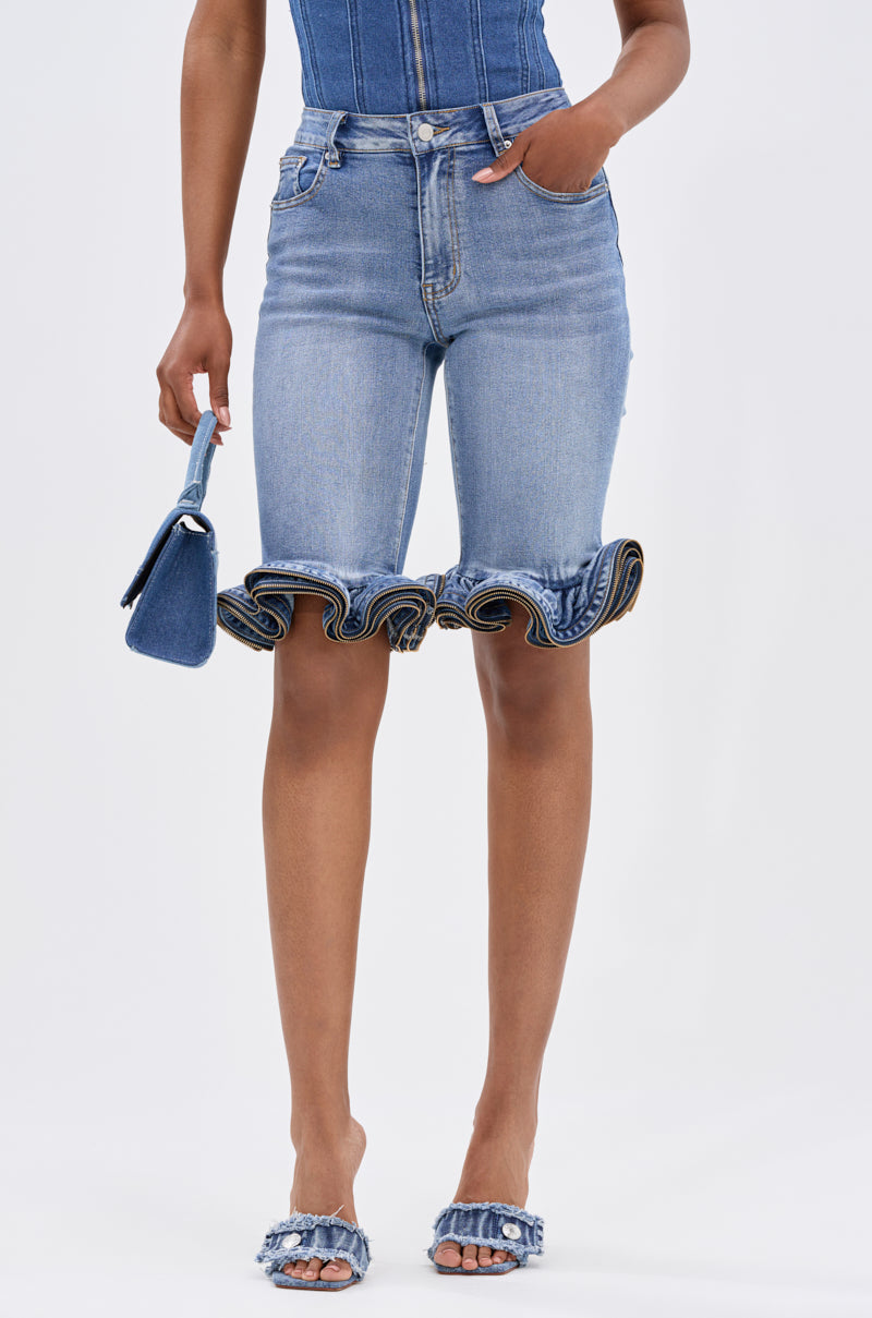 Wearing LONDON BERMUDA SHORT in light blue denim with ruffled hems, a person holds a small matching denim handbag and wears coordinating slide sandals with ruffle accents, set against a plain light background.