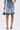 Wearing LONDON BERMUDA SHORT in light blue denim with ruffled hems, a person holds a small matching denim handbag and wears coordinating slide sandals with ruffle accents, set against a plain light background.