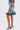 A woman models the LONDON BERMUDA SHORT—knee-length with ruffled details—paired with a blue top and heeled sandals, holding a small denim handbag. Side view highlights the shorts against a plain white background.