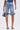 A woman stands facing away, wearing the LONDON BERMUDA SHORT with ruffled hems, a matching sleeveless top, denim high-heel shoes, and holding a small blue handbag.