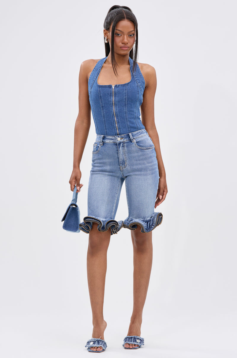 A woman poses against a plain background wearing the LONDON BERMUDA SHORT with ruffled hems, a fitted denim zip-up top, matching heeled sandals, and holding a small denim handbag.