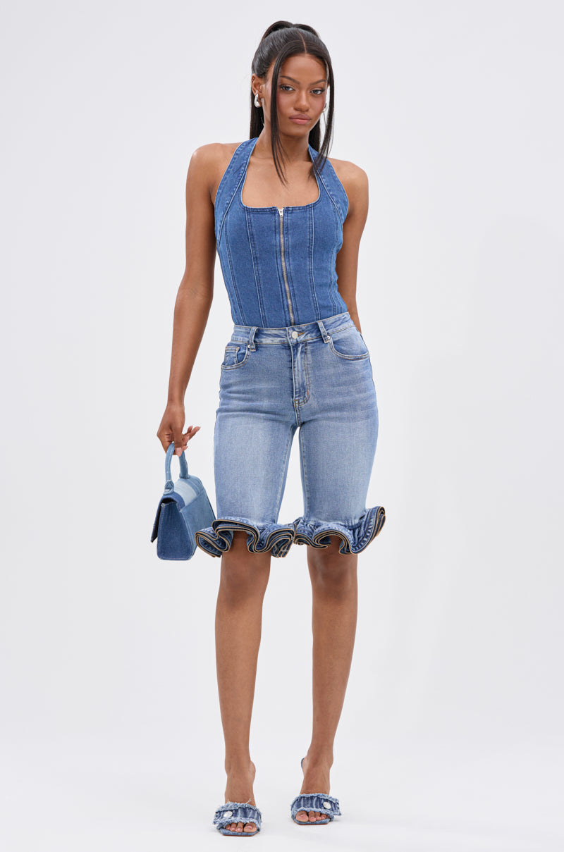A woman with straight hair and a neutral expression faces forward, wearing the LONDON BERMUDA SHORT featuring ruffled hems, a denim halter top, heeled sandals, and holding a blue handbag.