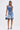 A woman with straight hair and a neutral expression faces forward, wearing the LONDON BERMUDA SHORT featuring ruffled hems, a denim halter top, heeled sandals, and holding a blue handbag.