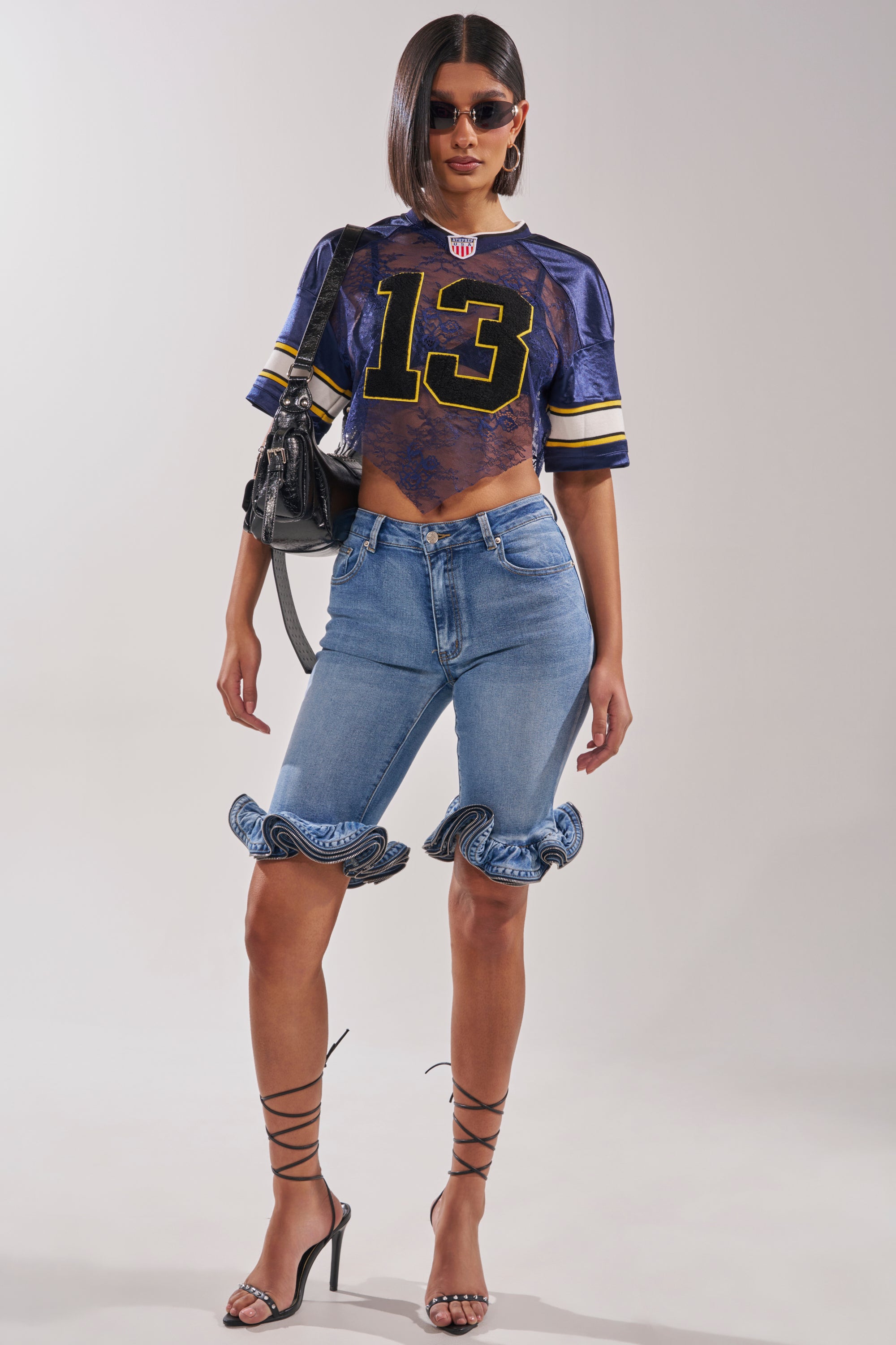 A woman wearing sunglasses, a cropped sports jersey with the number 13, LONDON BERMUDA SHORTS with ruffled hems, strappy black heels, and a black handbag poses against a plain background. 6