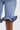 Close-up of a person wearing the LONDON BERMUDA SHORT, featuring blue denim with ruffled details, decorative zippers, and contrasting dark and gold trim, standing against a plain light background.