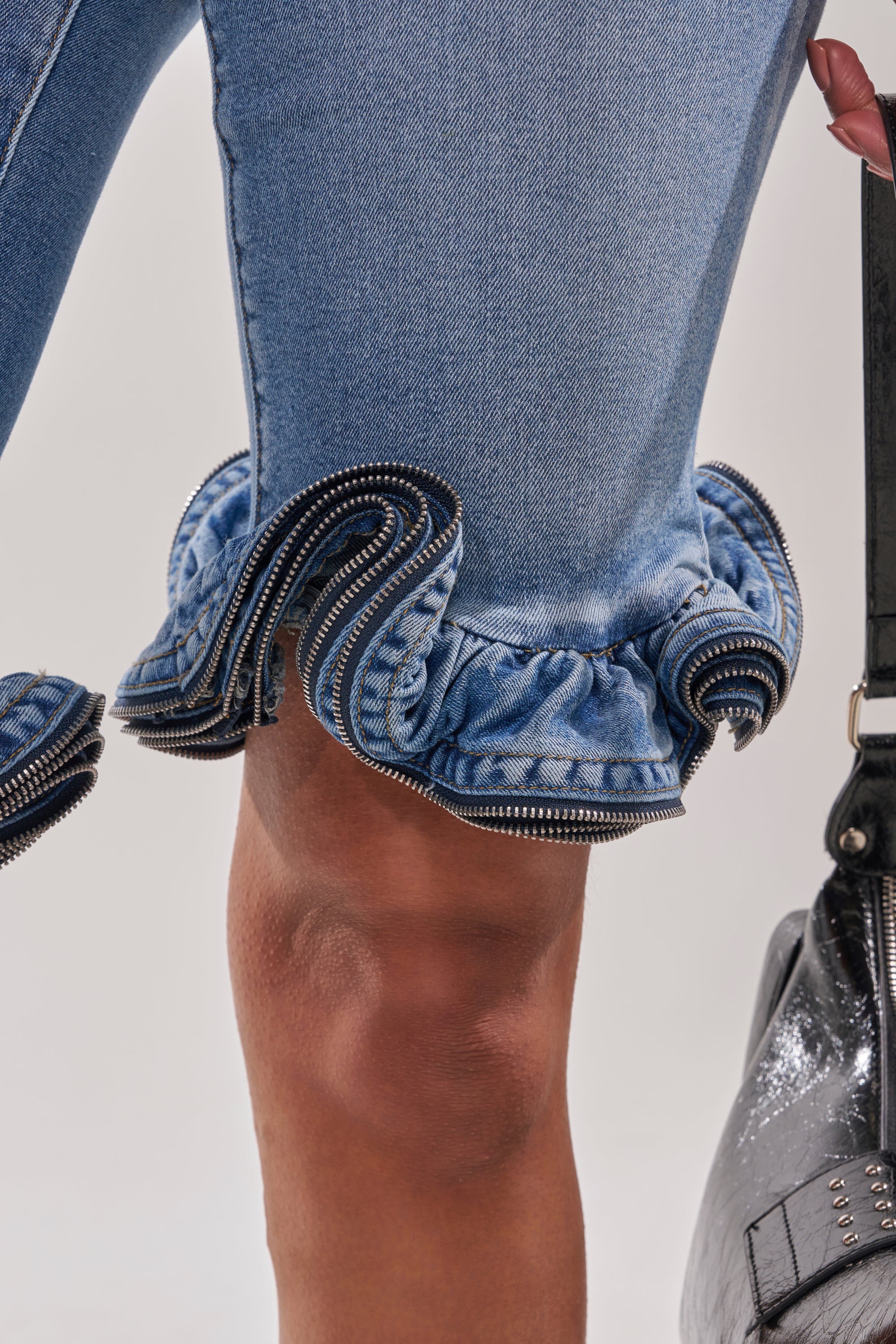 A close-up of a person in the LONDON BERMUDA SHORT, which features mid-rise denim and decorative ruffled zippers at the hem, holding a black leather bag with silver details. 8