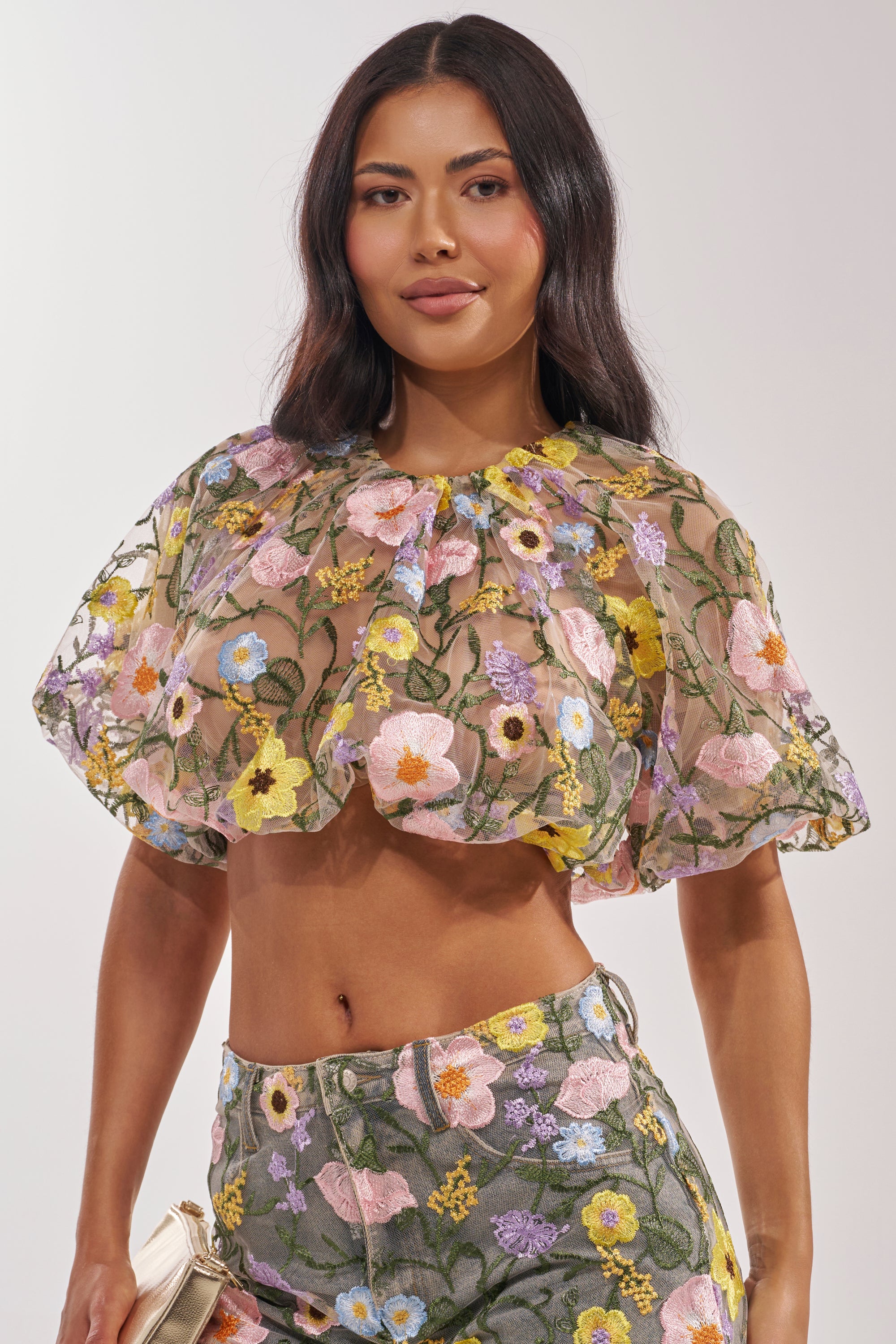 A woman in the GARDEN PARTY FLORAL BLOUSE, featuring sheer tulle, ruffles, and colorful floral embroidery, pairs it with high-waisted pants. She has long wavy dark hair, holds a small clutch, and stands against a plain light background. 1