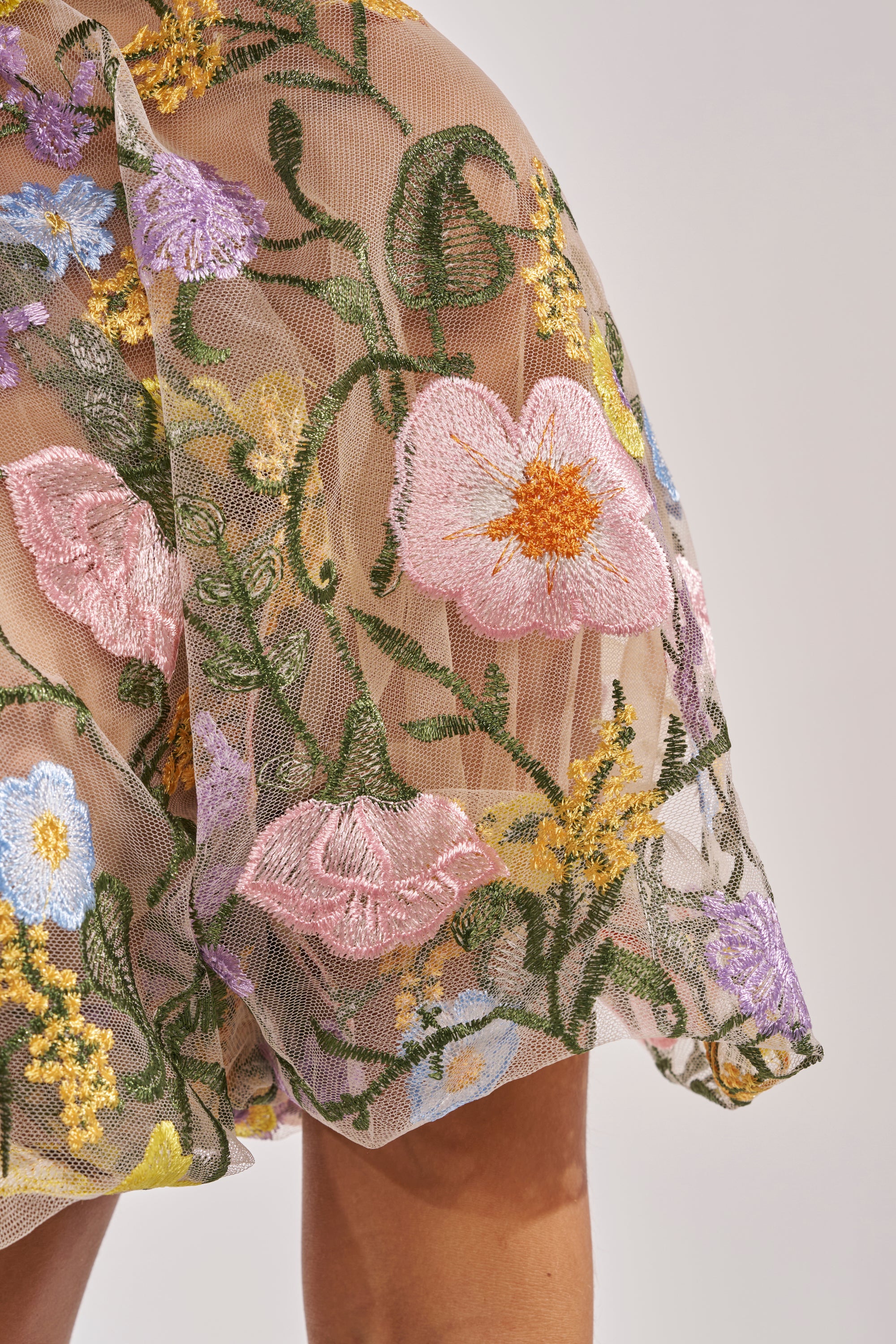 A close-up of the GARDEN PARTY FLORAL BLOUSE, a sheer tulle top adorned with colorful embroidered flowers, worn by a person with light brown skin. 10