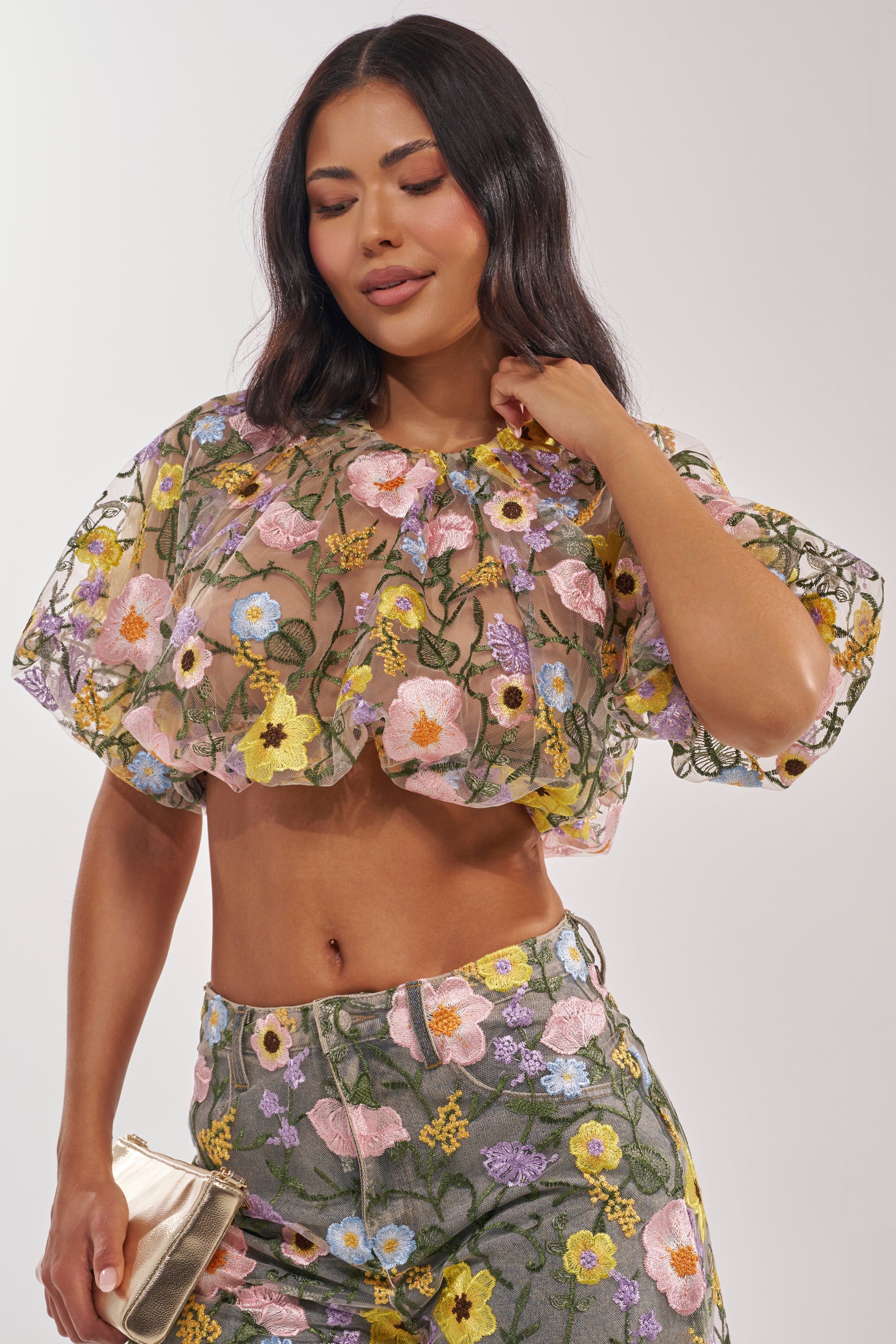 A woman in the GARDEN PARTY FLORAL BLOUSE and matching floral high-waisted pants holds a gold clutch and smiles with her eyes closed. 2