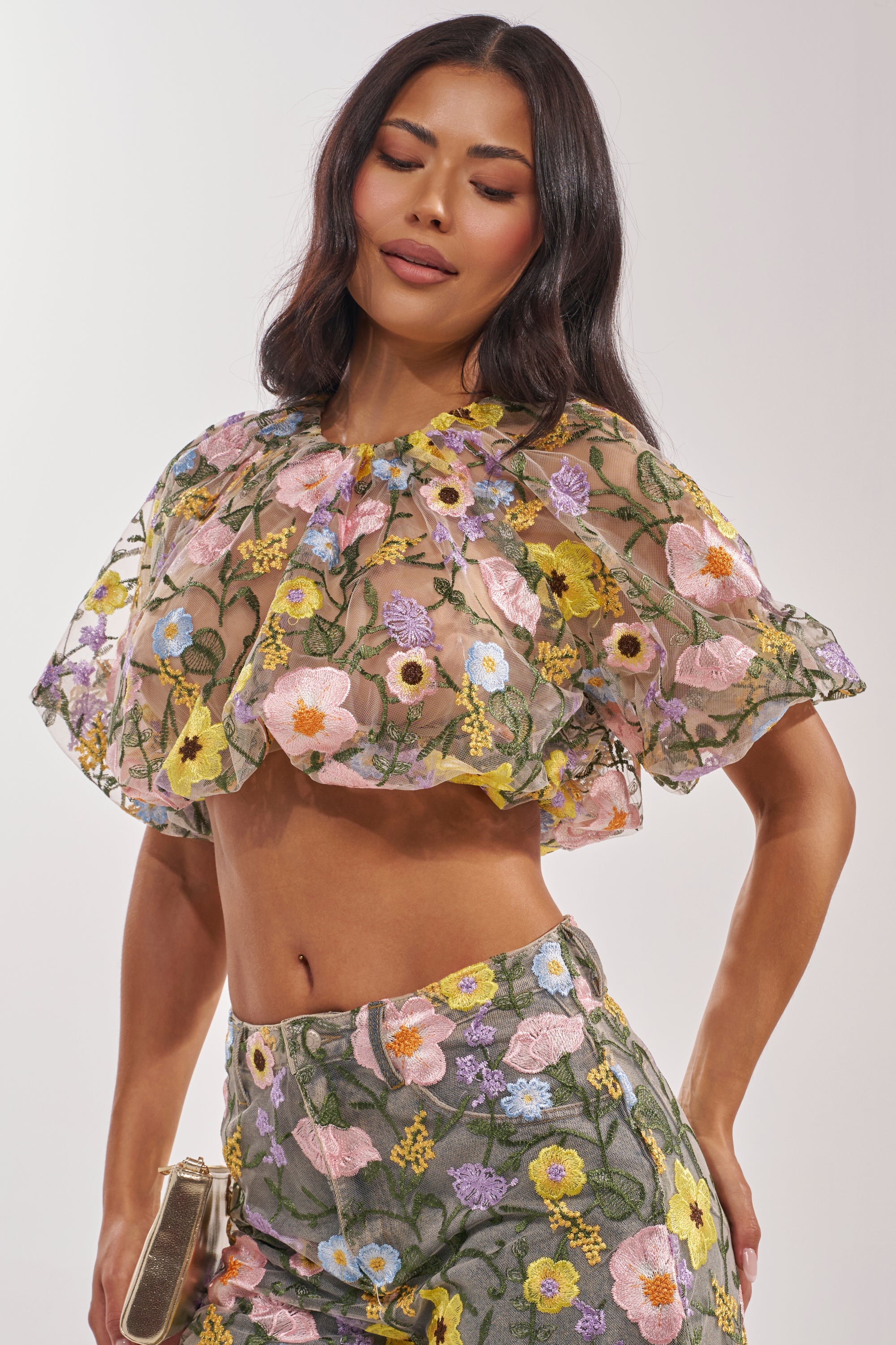 A woman wears the GARDEN PARTY FLORAL BLOUSE with matching high-waisted pants, featuring vibrant pink, yellow, and purple embroidery—perfect for a chic garden party. She accessorizes with a small clutch and soft waves in her hair. 3