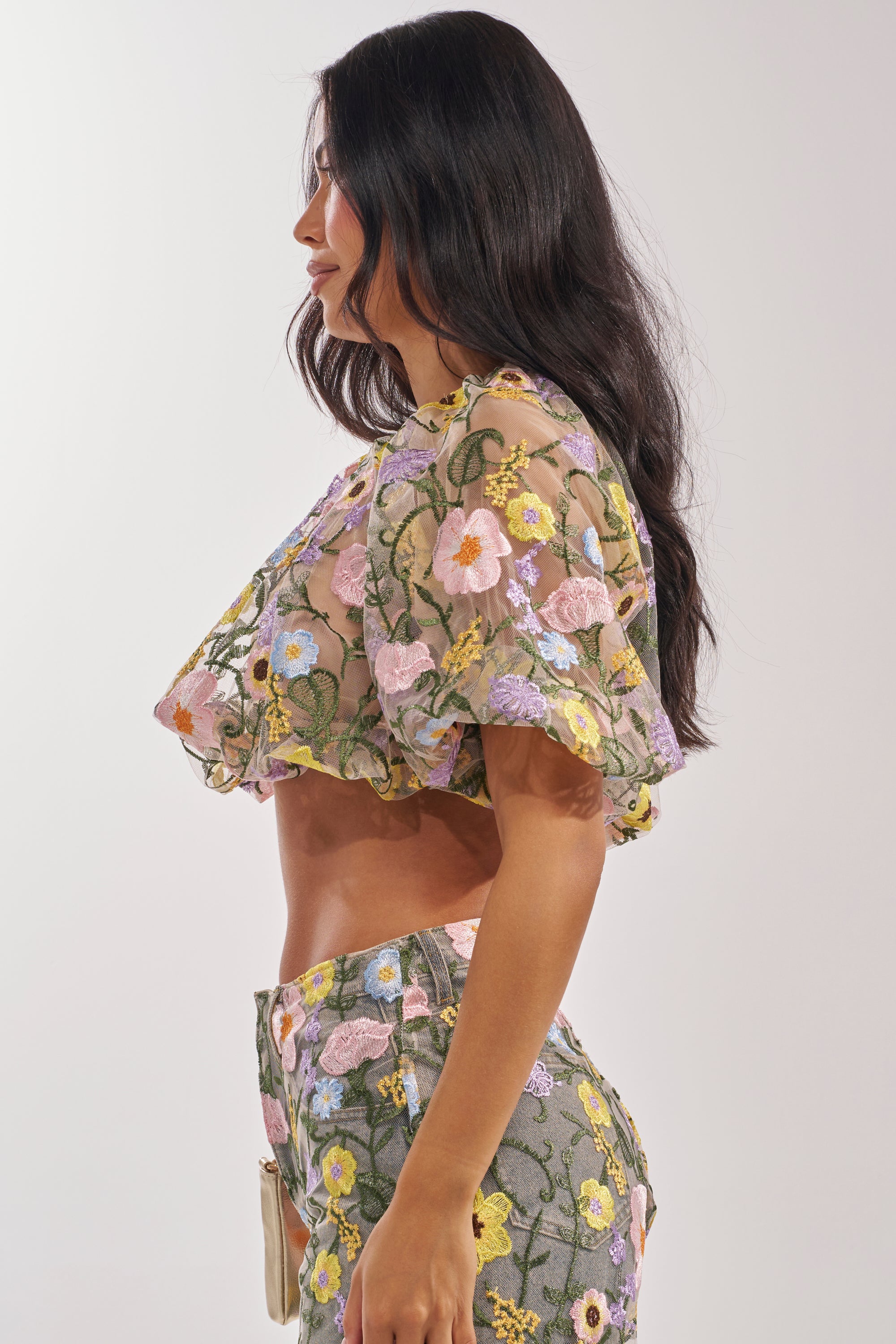 A woman with long dark hair wears the GARDEN PARTY FLORAL BLOUSE, a sheer tulle top adorned with colorful floral embroidery, standing in profile against a plain light background. 4