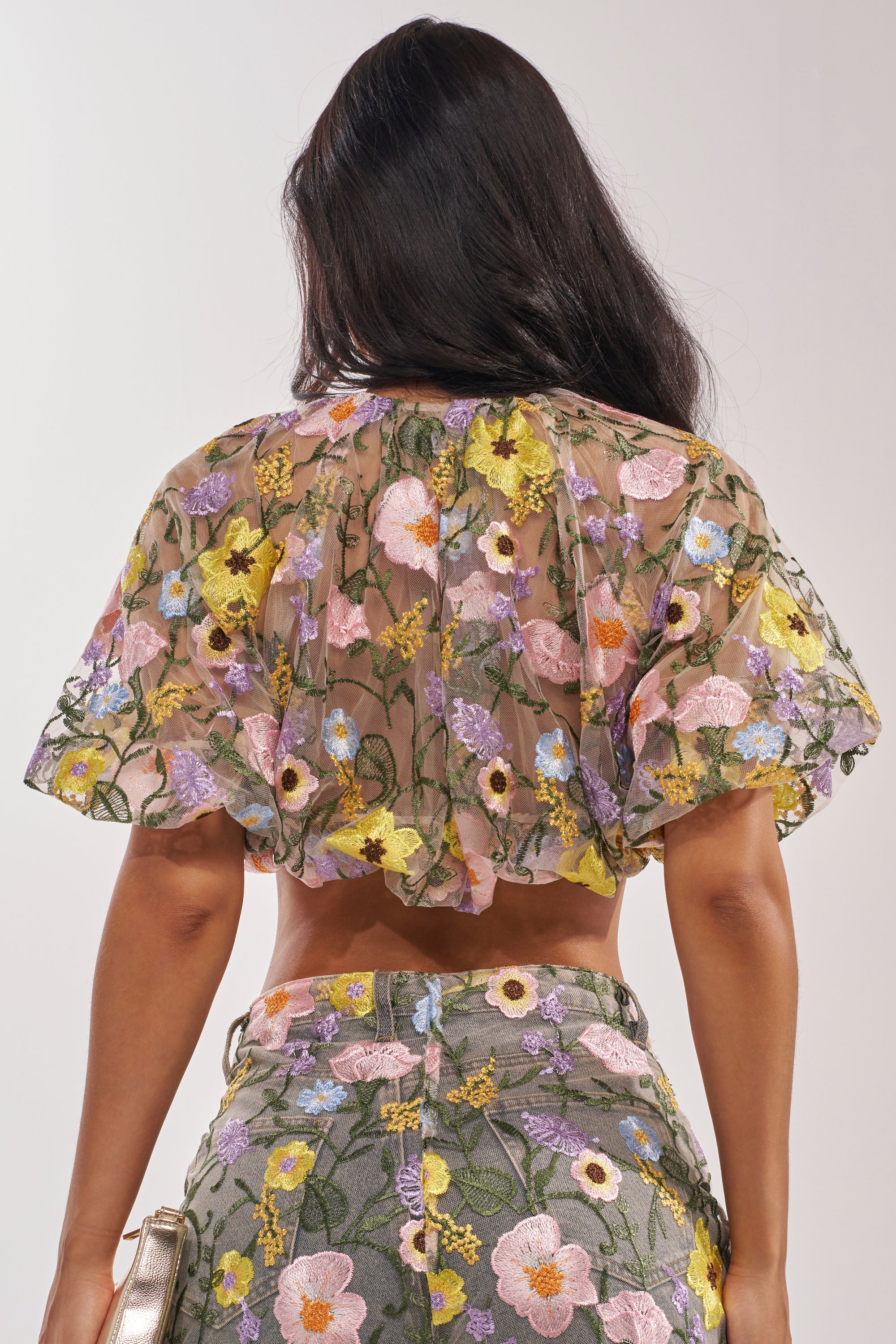 A woman with long dark hair faces away from the camera, showing the back of the GARDEN PARTY FLORAL BLOUSE—a sheer tulle top decorated with colorful embroidered flowers, paired with matching high-waisted pants. 5
